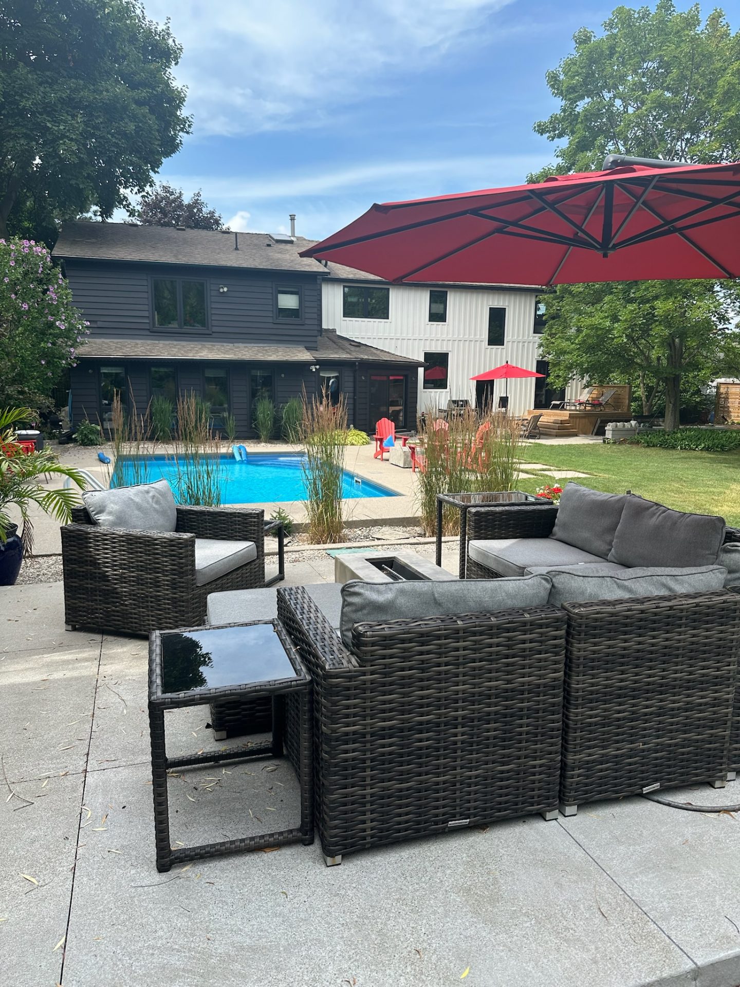 A modern backyard features a pool, wicker furniture, and red umbrellas. A two-story house and green trees create a serene atmosphere.
