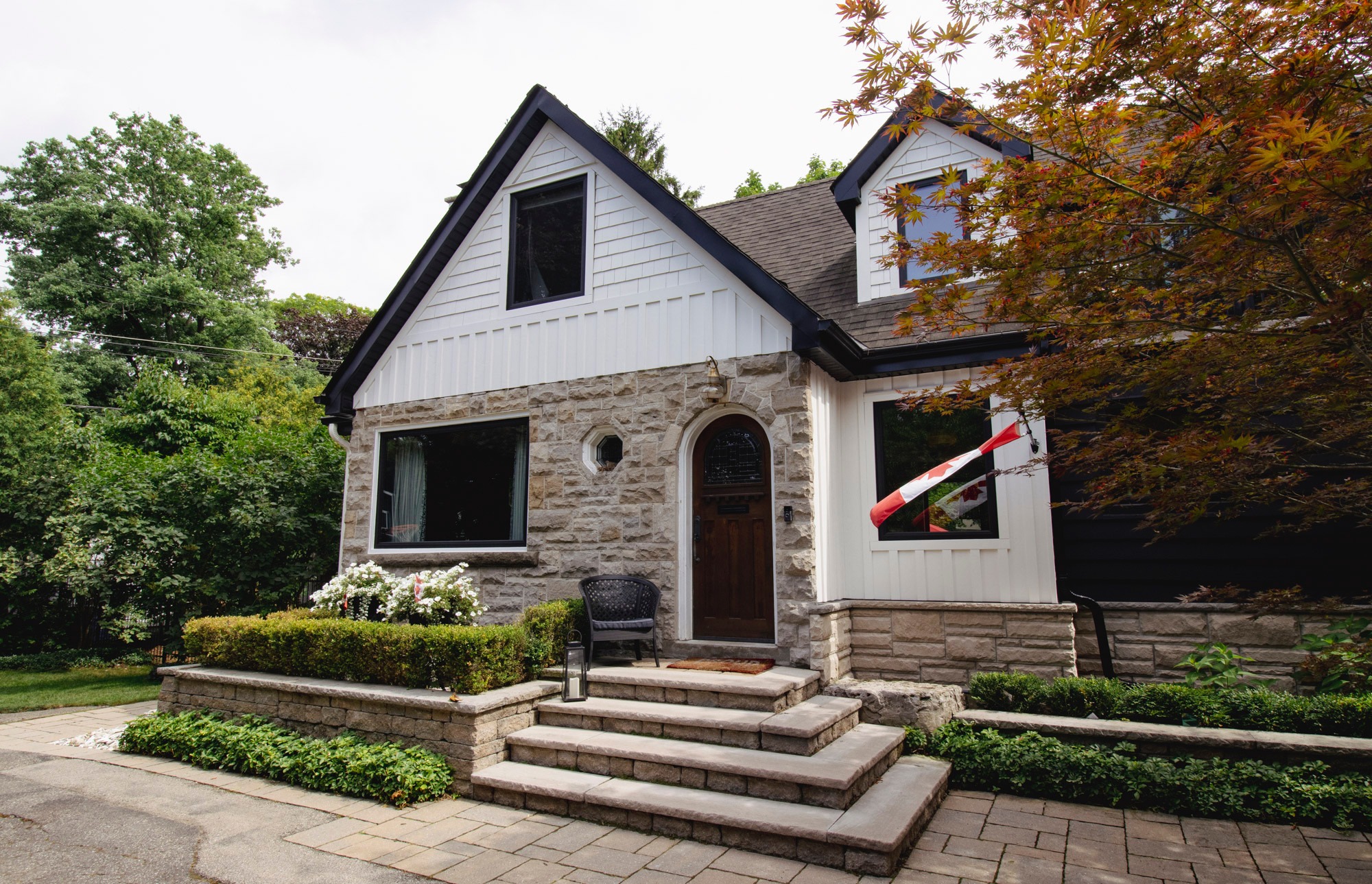 Charming stone house with a Canadian flag, surrounded by lush greenery and a neatly landscaped garden. Steps lead to a wooden door.