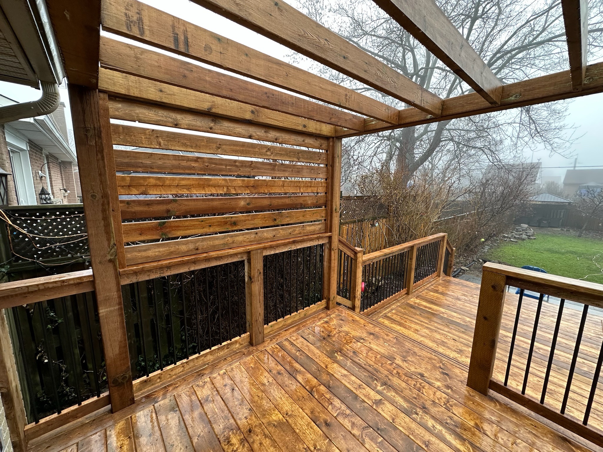 A wooden deck with a pergola overlooks a foggy backyard garden, featuring bare trees and a small shed in the background.