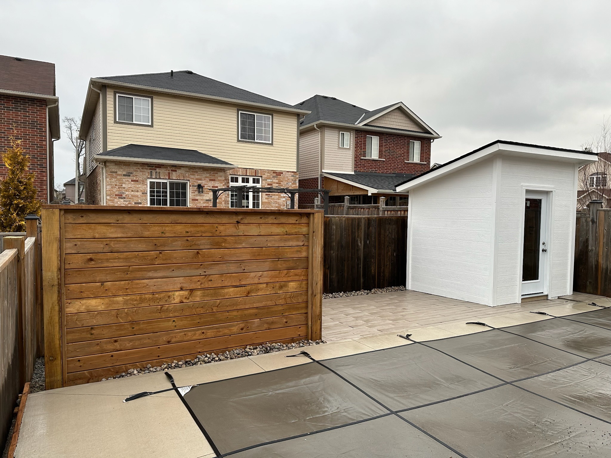 Fenced backyard with white shed, tiled patio, and covered pool. Two neighboring brick houses are visible in the background under overcast skies.