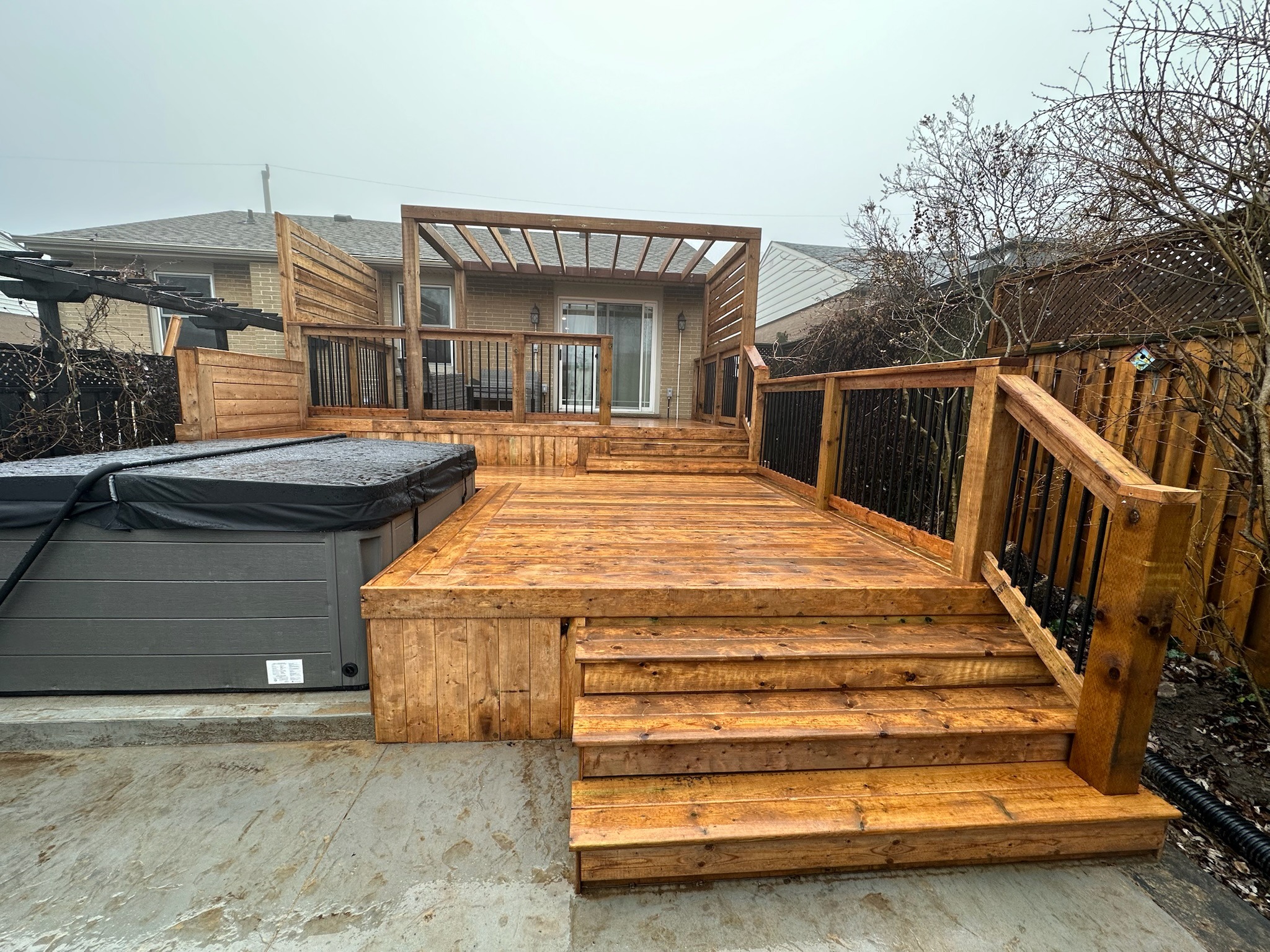 A wooden deck with steps, hot tub, and pergola is in a backyard. The scene is overcast, with bare trees and fences.