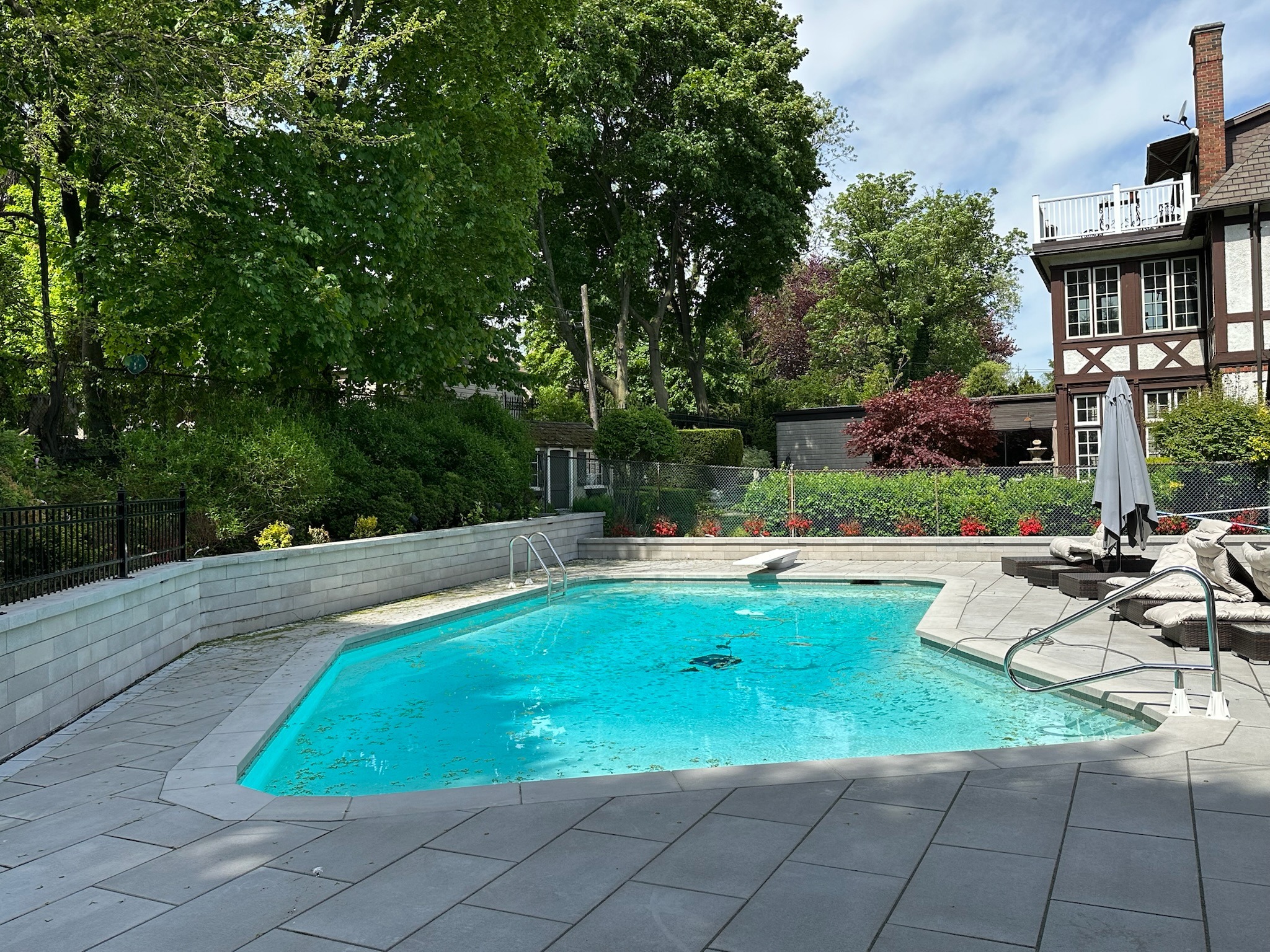 A picturesque backyard view features a clear blue swimming pool, surrounded by lush greenery and adjacent to a Tudor-style house.