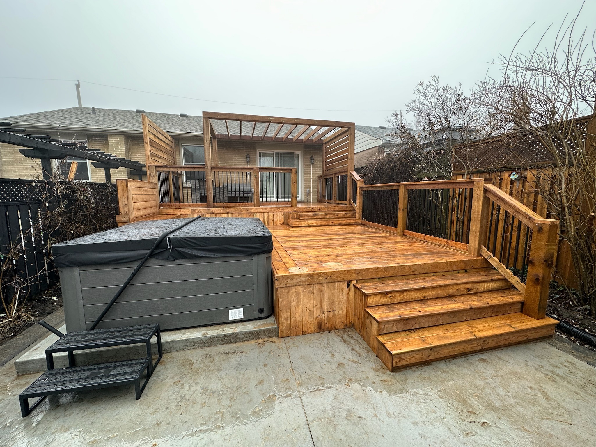 Wooden deck with steps, a hot tub, and a pergola in a backyard. Leafless trees and a residential building surround the area.