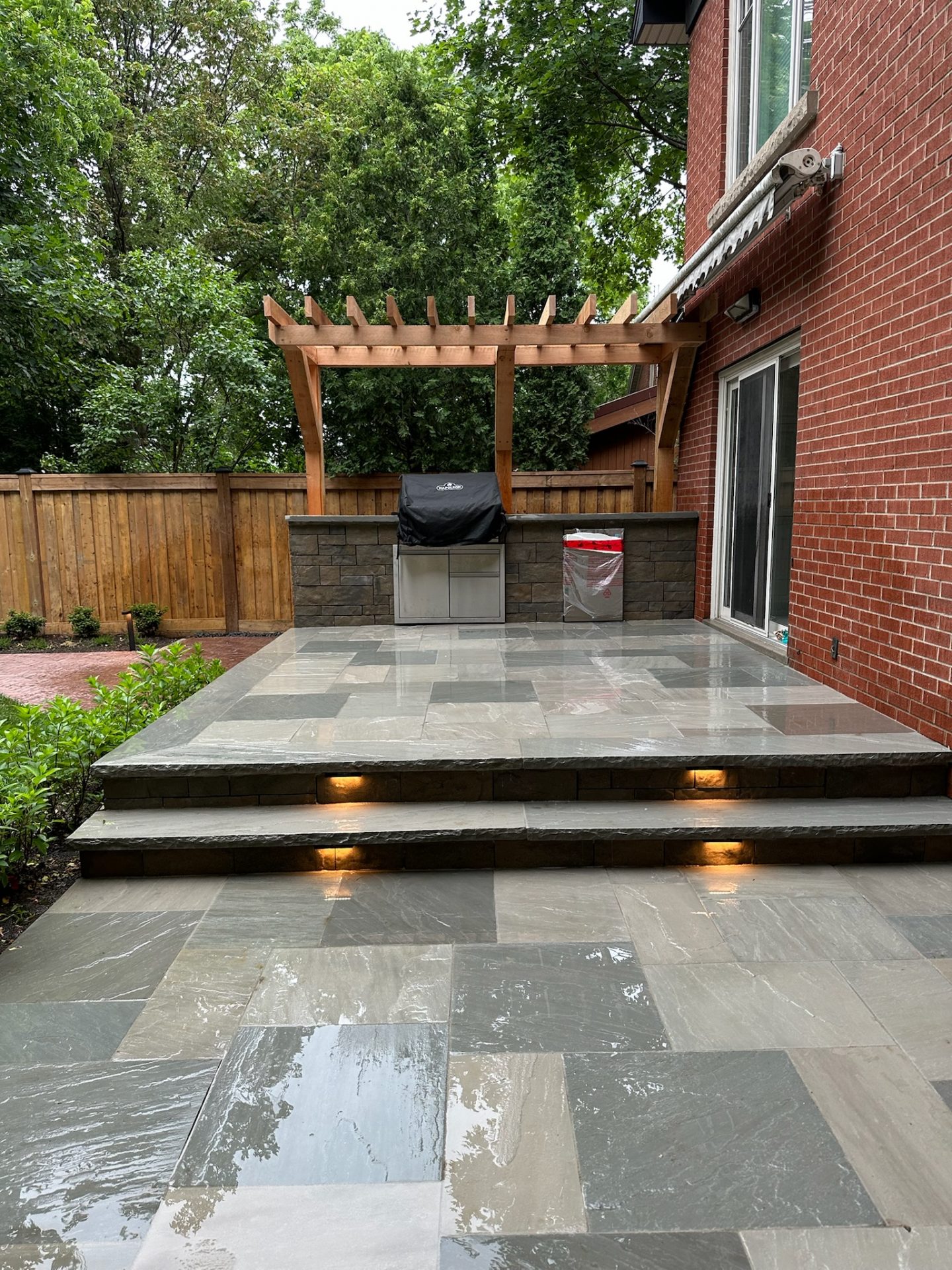 Stone patio with illuminated steps, a pergola over an outdoor grill, surrounded by trees and a brick house. Wooden fence in background.