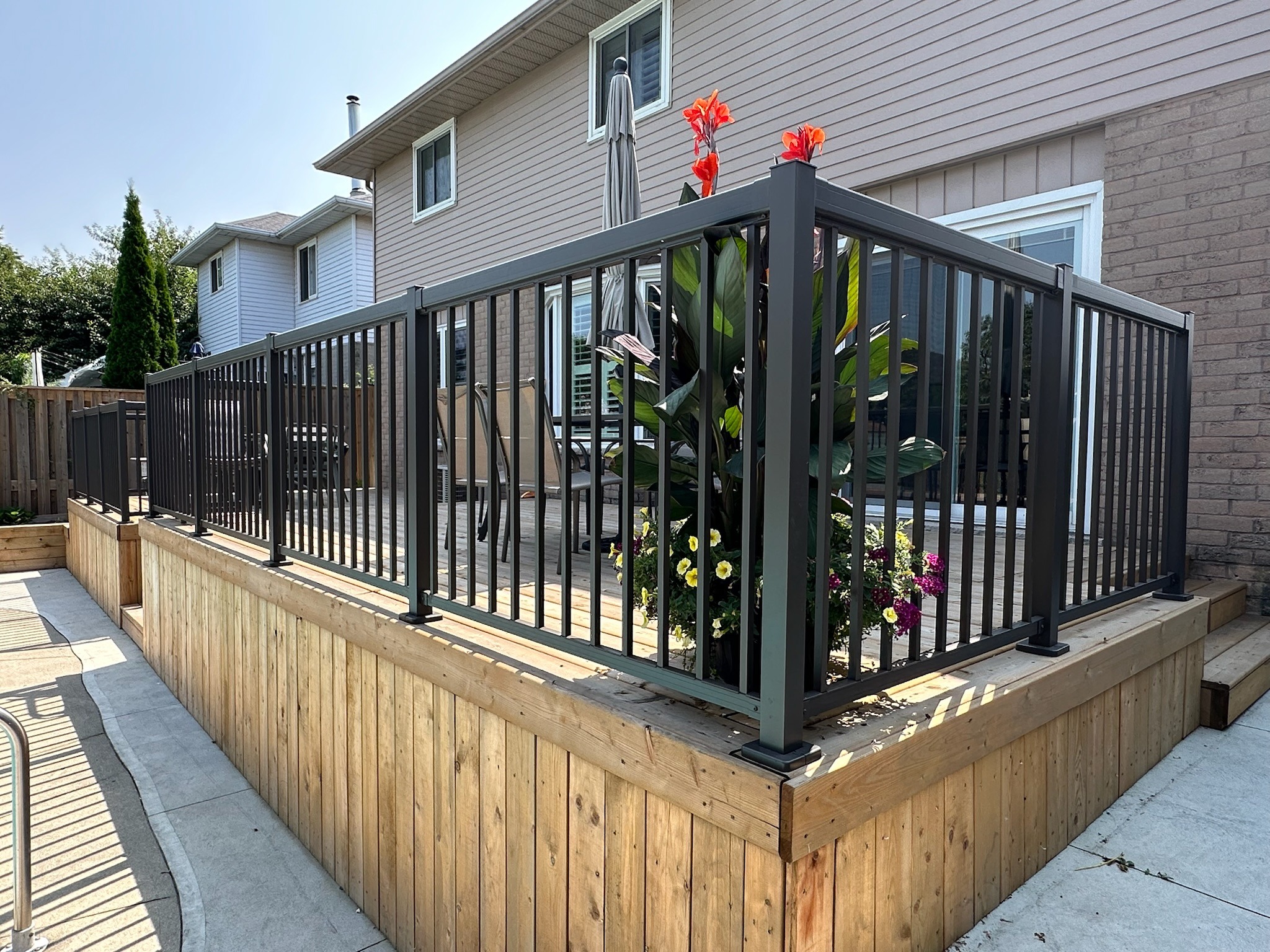 A wooden deck with modern railing and vibrant flowers is attached to a house in a suburban neighborhood, under a clear sky.