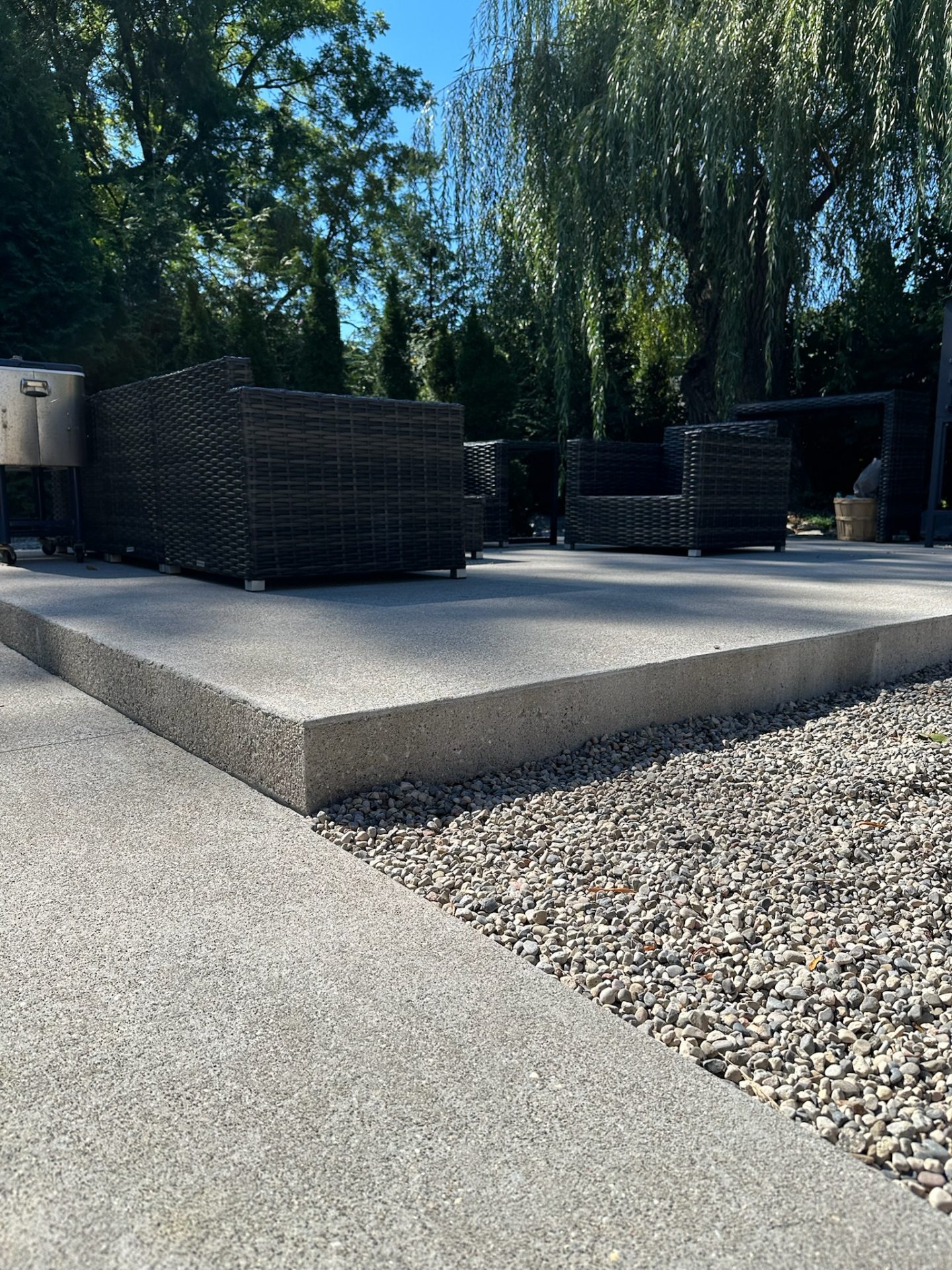 Outdoor patio scene with wicker furniture, surrounded by lush trees and gravel, under bright daylight. Peaceful and inviting atmosphere.