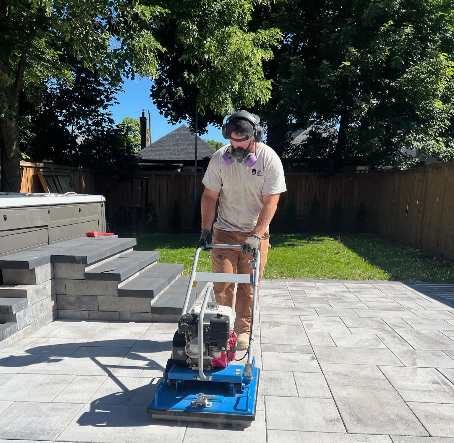 A person operates a plate compactor on a tiled patio in a backyard, wearing protective gear. Trees and a wooden fence are visible.