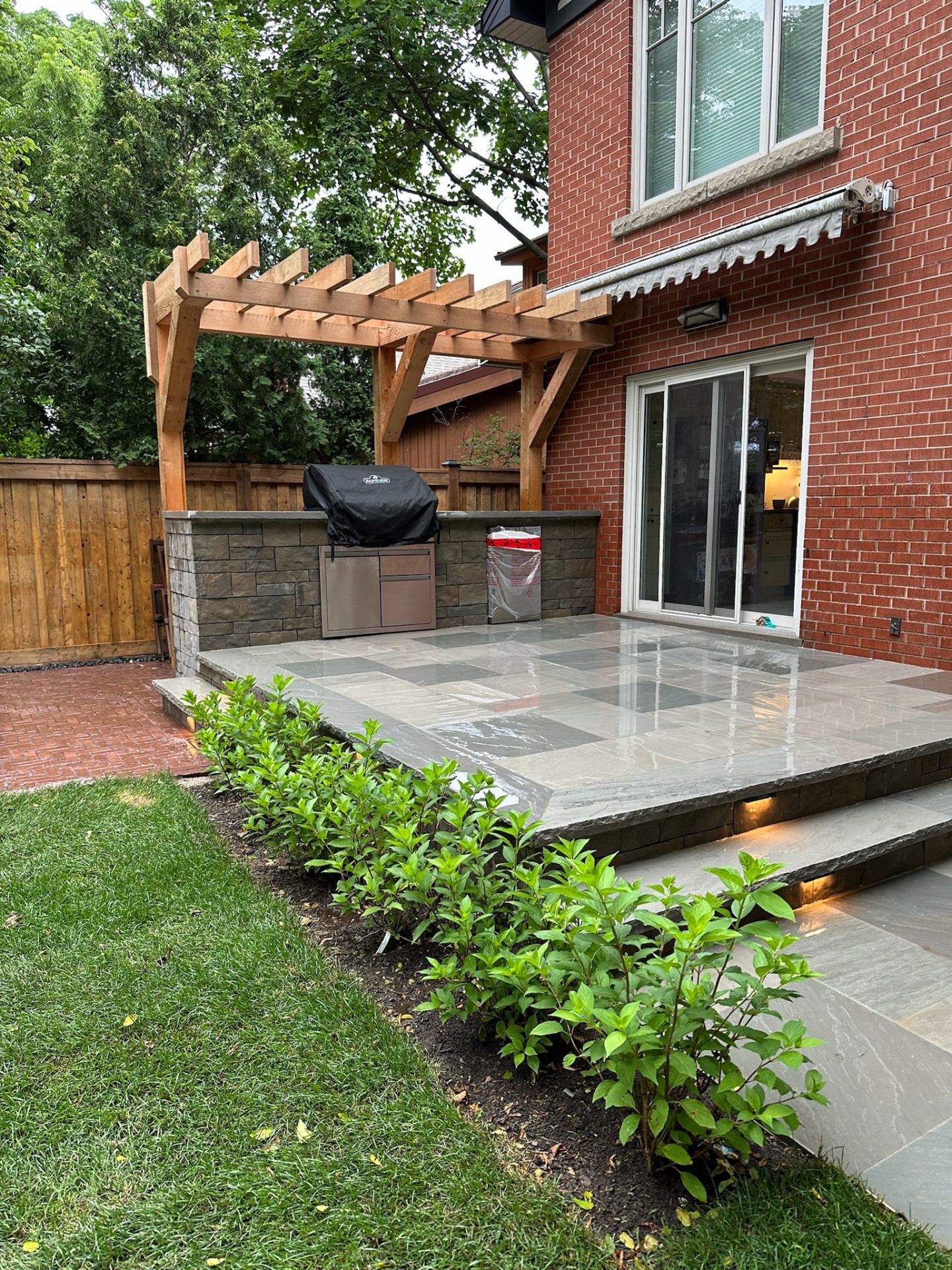 Brick house patio with wooden pergola, stone grill area, sliding glass door, lush hedge, and greenery. Modern outdoor living space covered by trees.