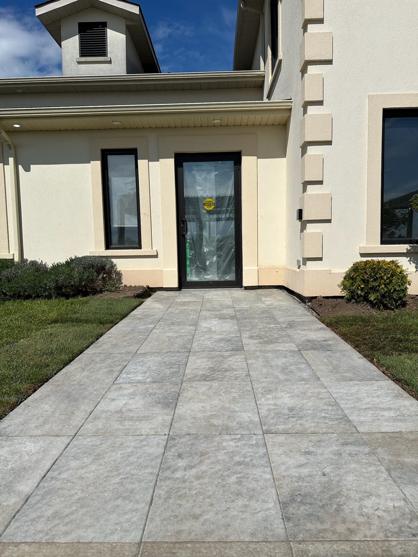 Modern building facade featuring a tiled walkway, beige walls, and large windows under a clear blue sky. No recognizable landmarks or historical buildings.