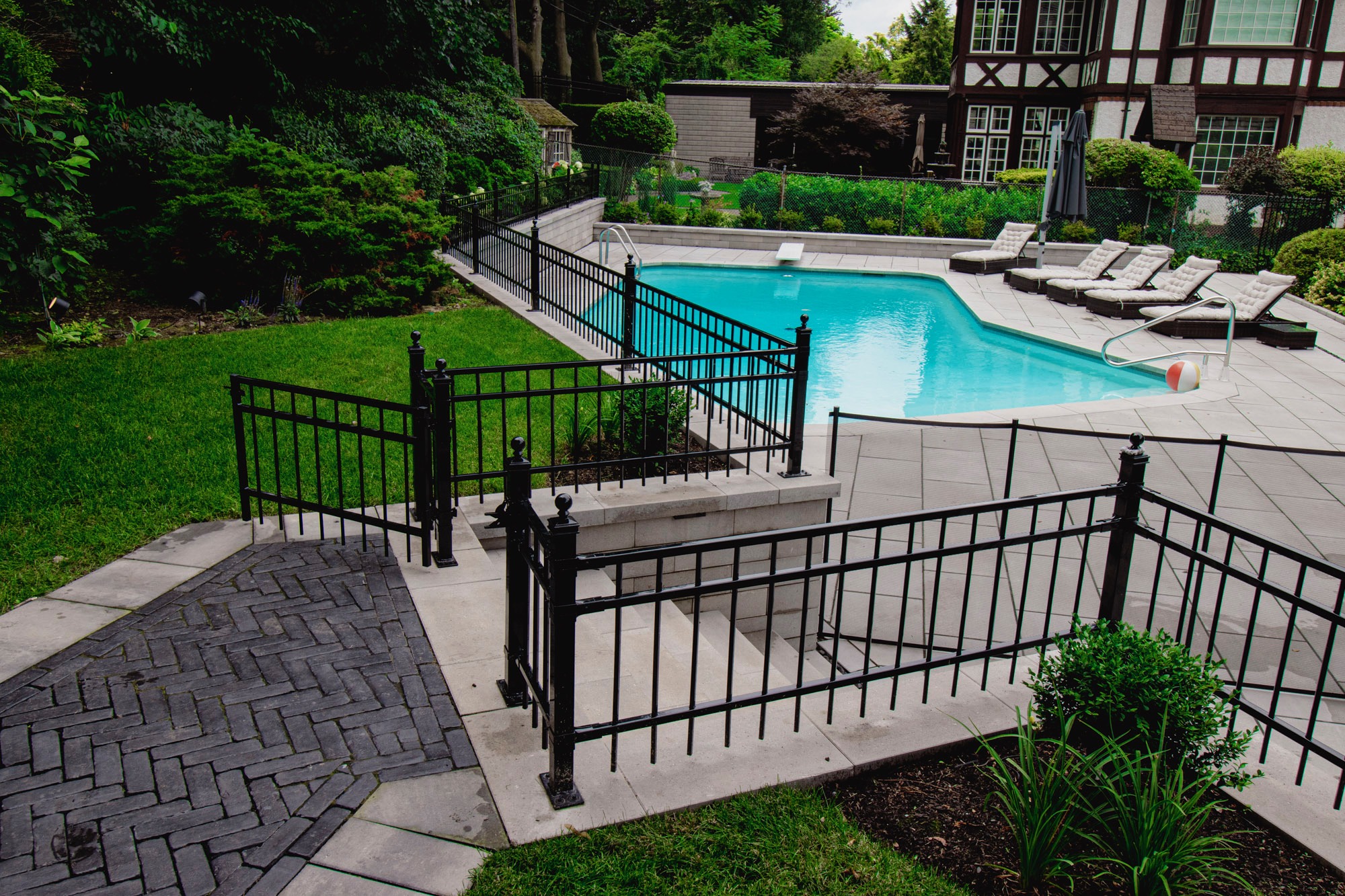 A serene backyard features a clean, inviting swimming pool surrounded by elegant black fencing, lush greenery, and a classic house exterior.