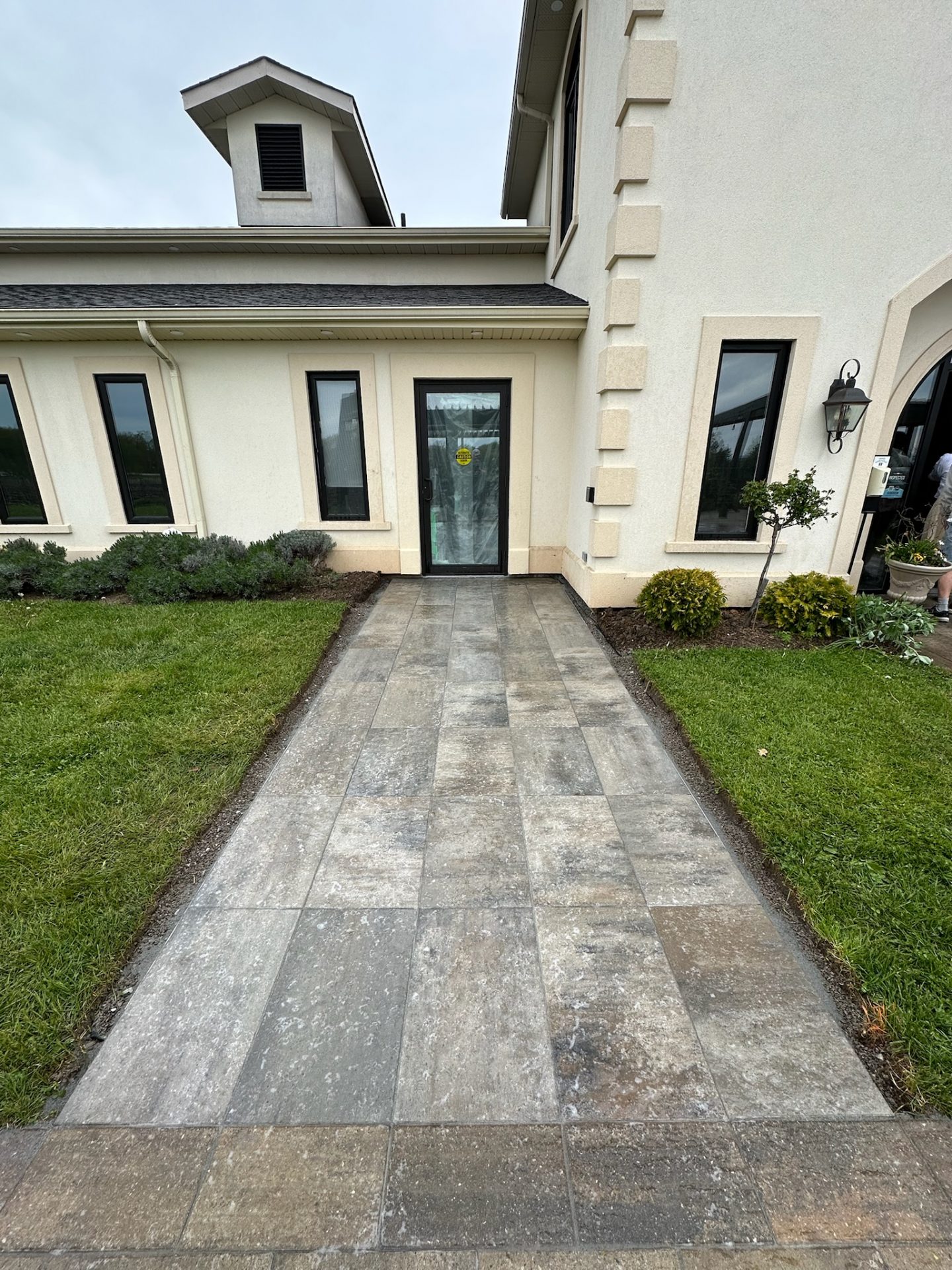 A modern building with tall windows and a stone pathway is flanked by neatly cut grass and small shrubs, under an overcast sky.
