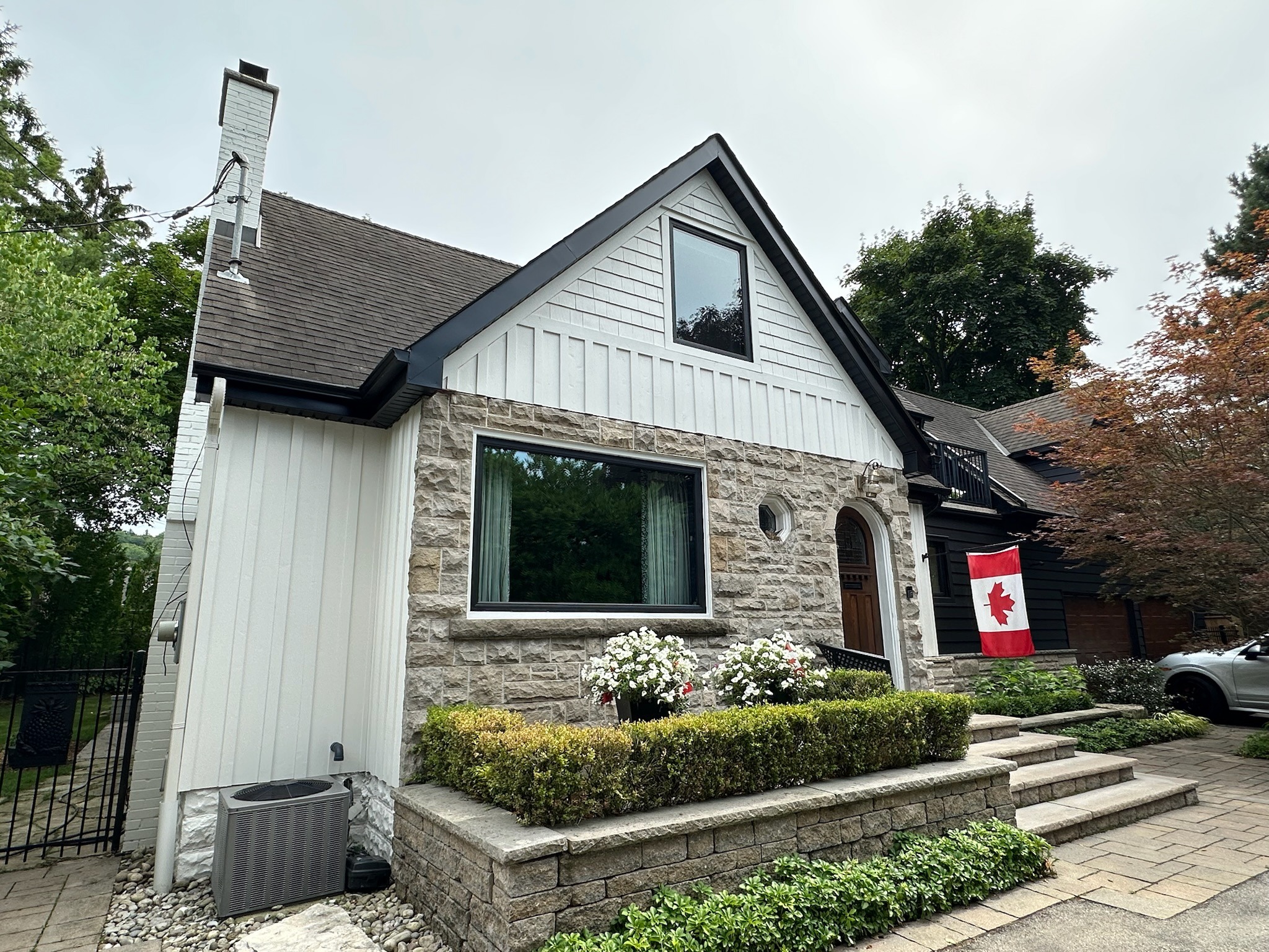 A charming house with stone facade, Canadian flag, and well-maintained garden, surrounded by trees. A gray SUV is parked nearby.