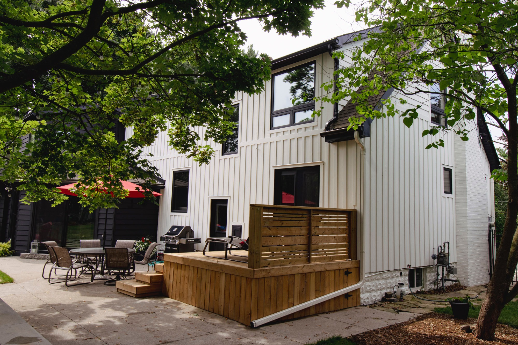 Modern two-story house with white exterior, patio furniture, and red umbrella. Wooden deck extends into the garden, surrounded by trees.