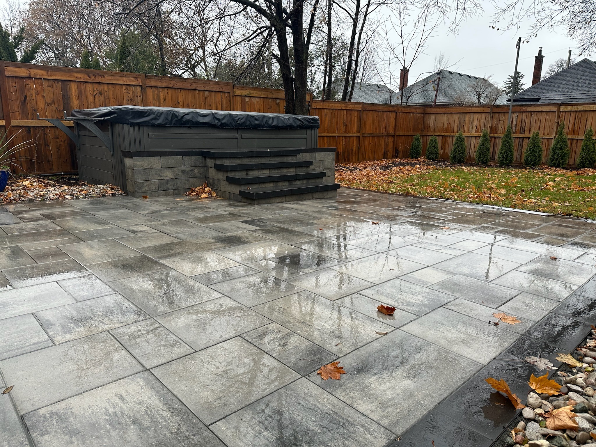 A backyard features wet stone tiles, a covered hot tub, wooden fence, trees, and scattered autumn leaves under an overcast sky.