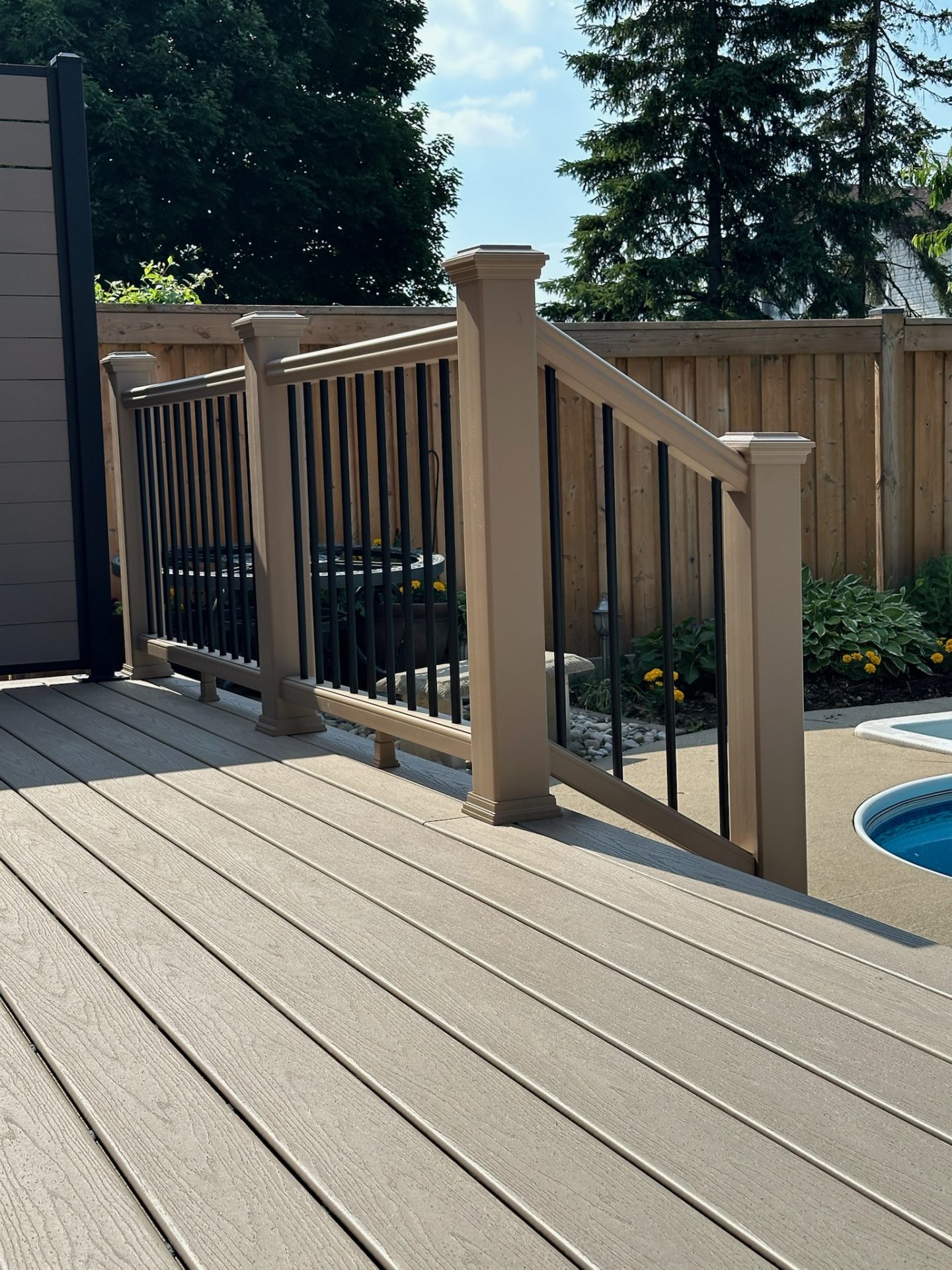 A wooden deck with railing overlooks a backyard pool, surrounded by a wooden fence and lush greenery. No landmarks or buildings visible.