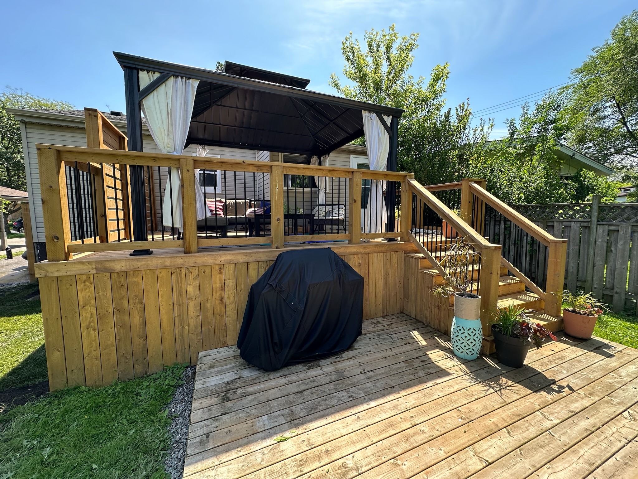 Wooden patio with stairs leading to a covered gazebo, surrounded by potted plants and trees under a clear blue sky.