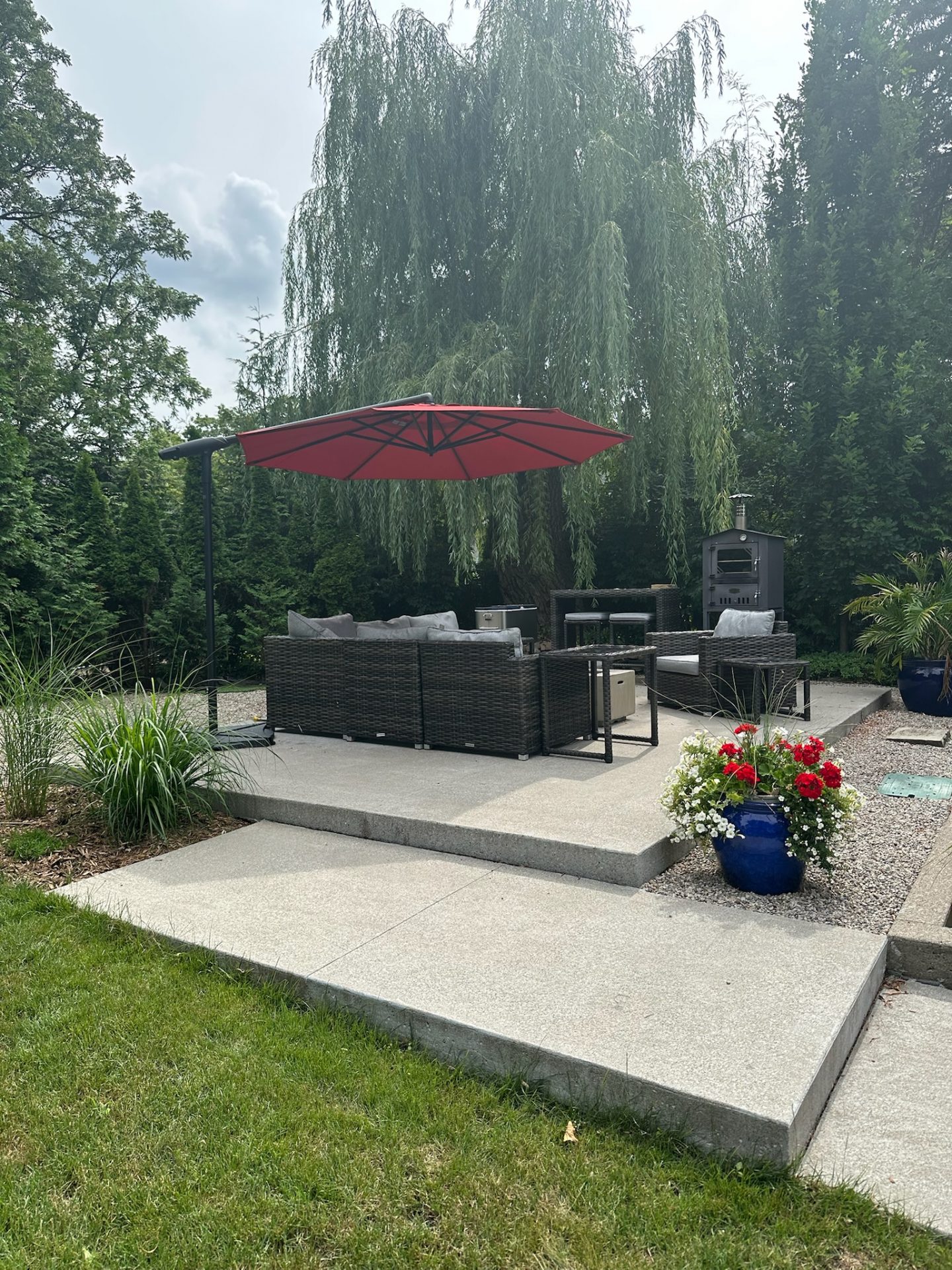 A modern patio with wicker furniture, red umbrella, and decorative plants. Surrounded by trees, with a serene garden ambiance.