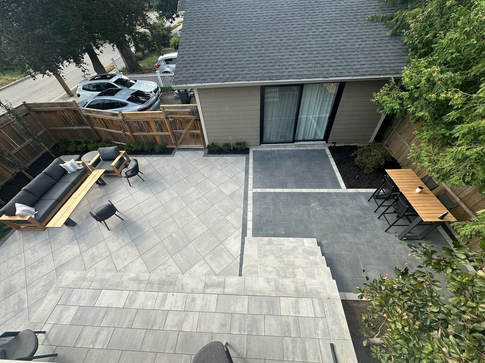 Modern backyard patio with gray tiles, outdoor seating, and wooden fence. Small garden, trees, and two parked cars visible near the entrance.