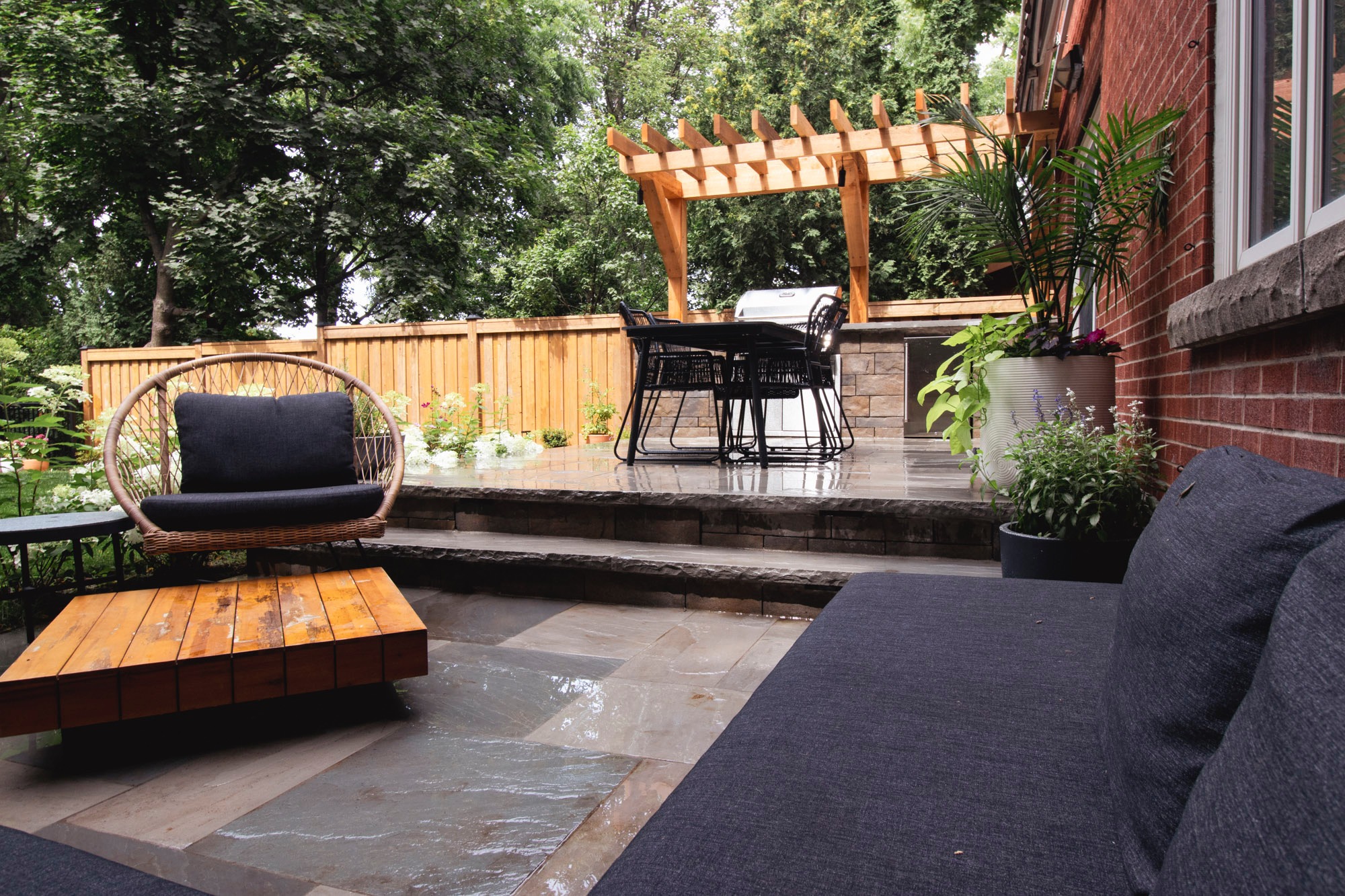 A modern patio features wooden pergola, wicker furniture, and potted plants, surrounded by lush greenery and a wooden fence in a serene outdoor space.