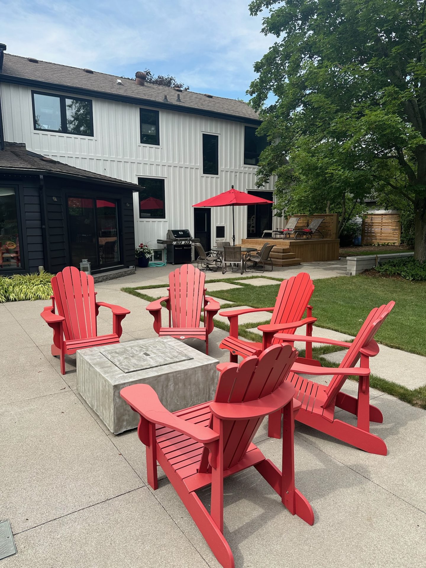 A modern backyard features red chairs, a concrete table, and patio with umbrella. White house exterior and green tree in the background.