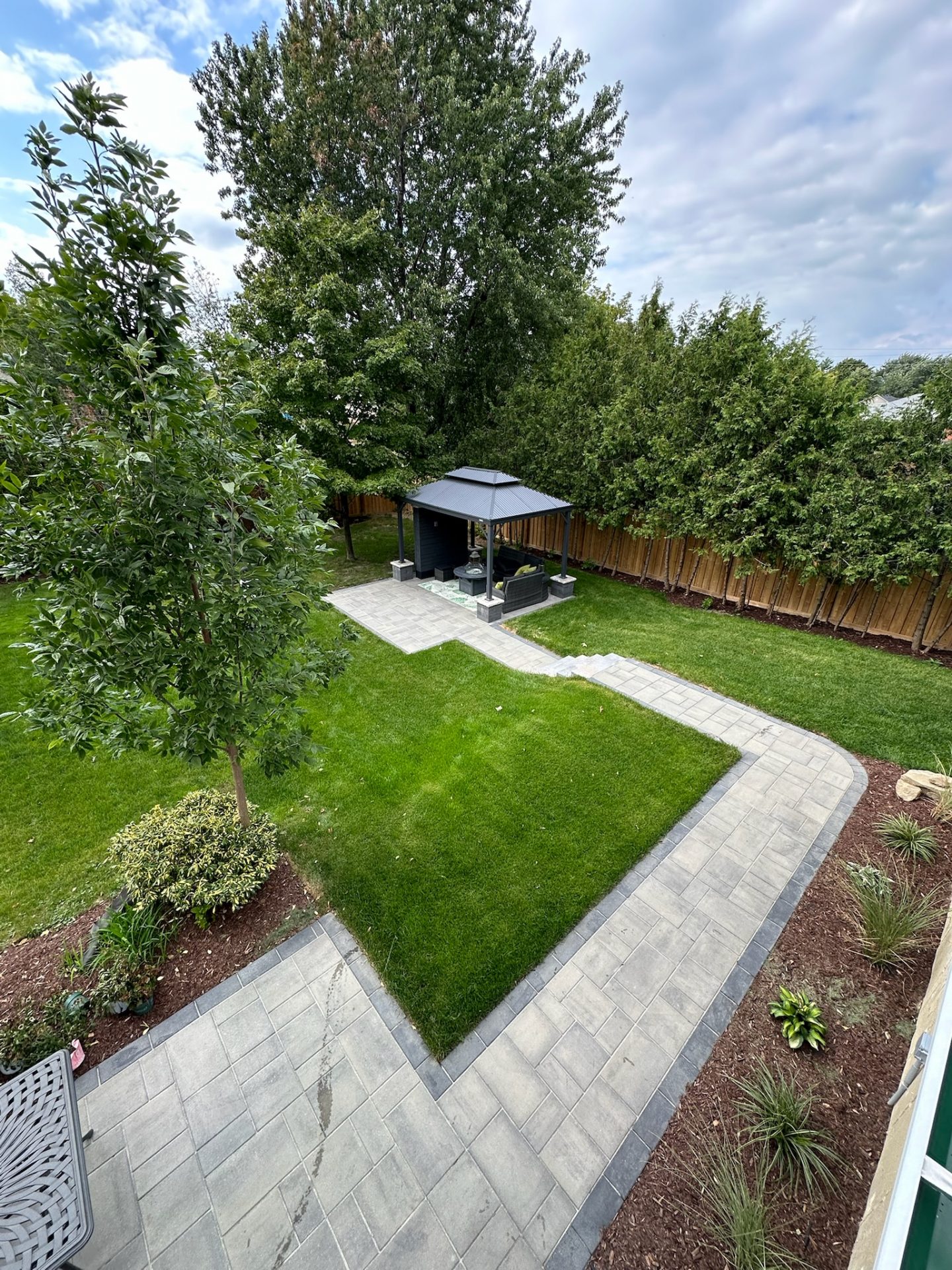 A neatly landscaped backyard features a paved walkway, green grass, and a gazebo under tall trees, all enclosed by a wooden fence.