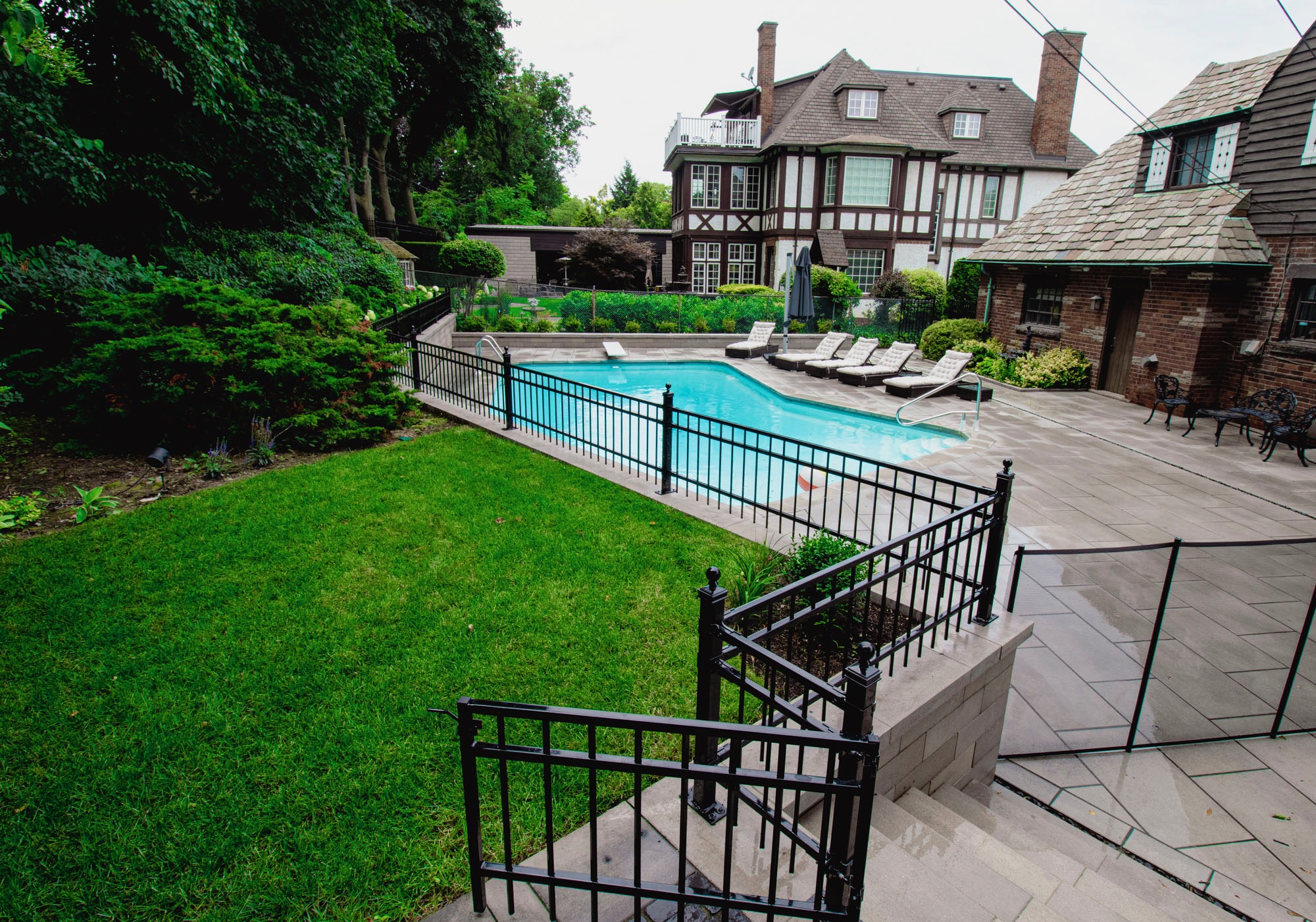 A Tudor-style house with a lush garden, fenced swimming pool, lawn chairs, and stone patio, set amidst surrounding greenery.