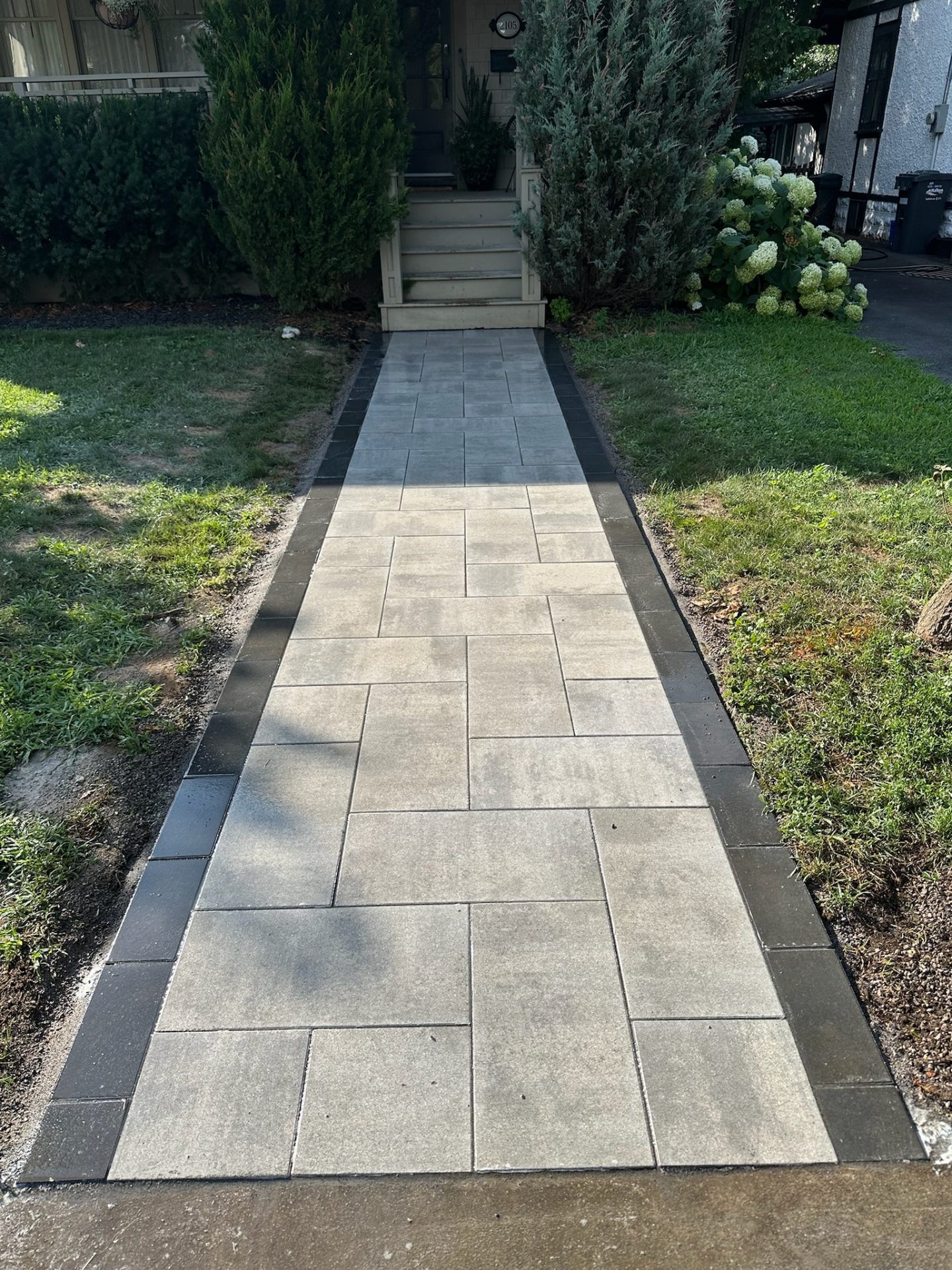 A tiled walkway leads to a porch with steps, surrounded by lush greenery and hydrangeas, in a peaceful residential setting.