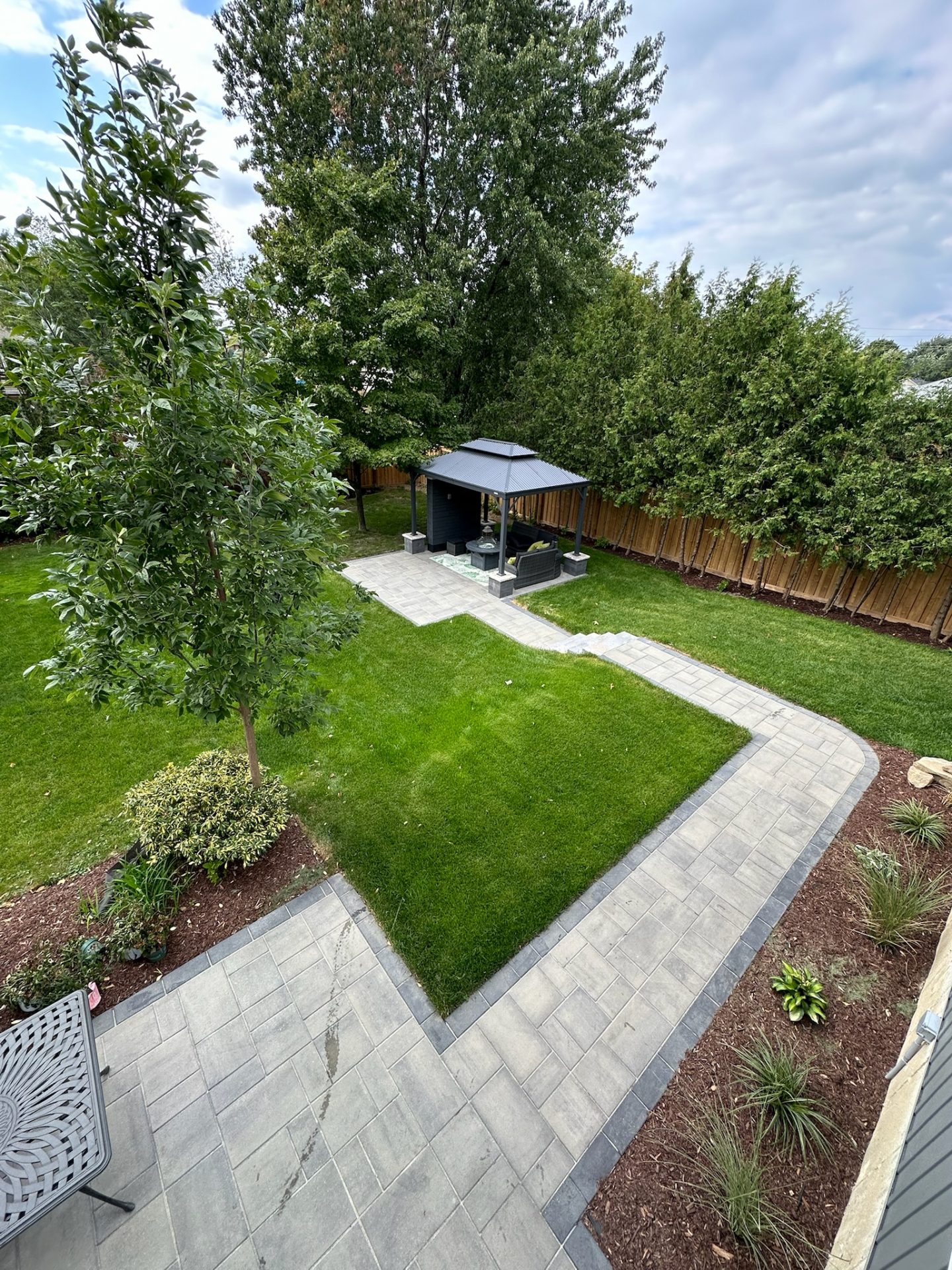 Aerial view of a backyard with a paved patio, lush lawn, garden beds, and a gazebo surrounded by trees and hedges under a cloudy sky.