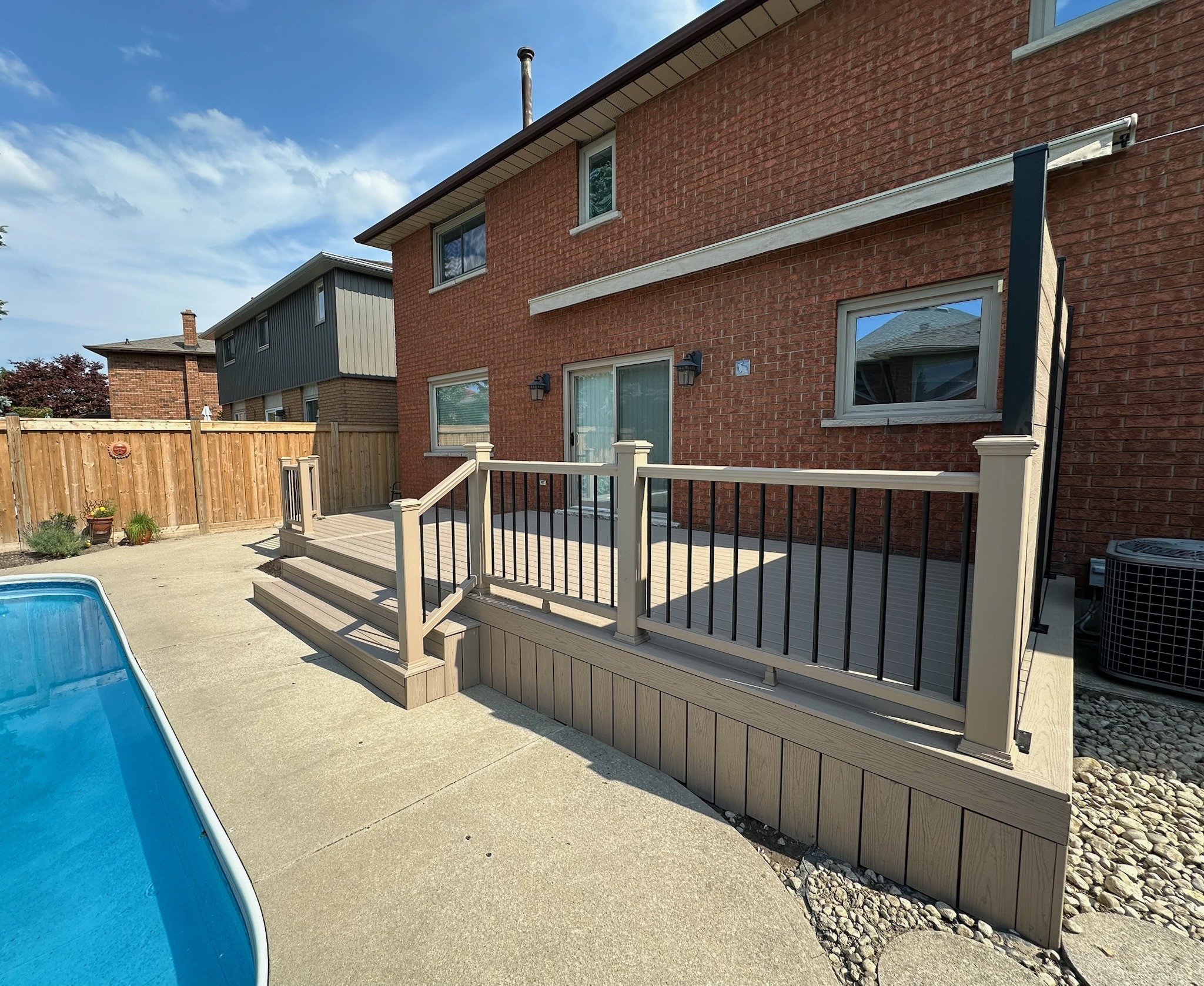 A backyard features a brick house, elevated deck, pool, and wooden fence under a partly cloudy sky. No landmarks or historical buildings visible.