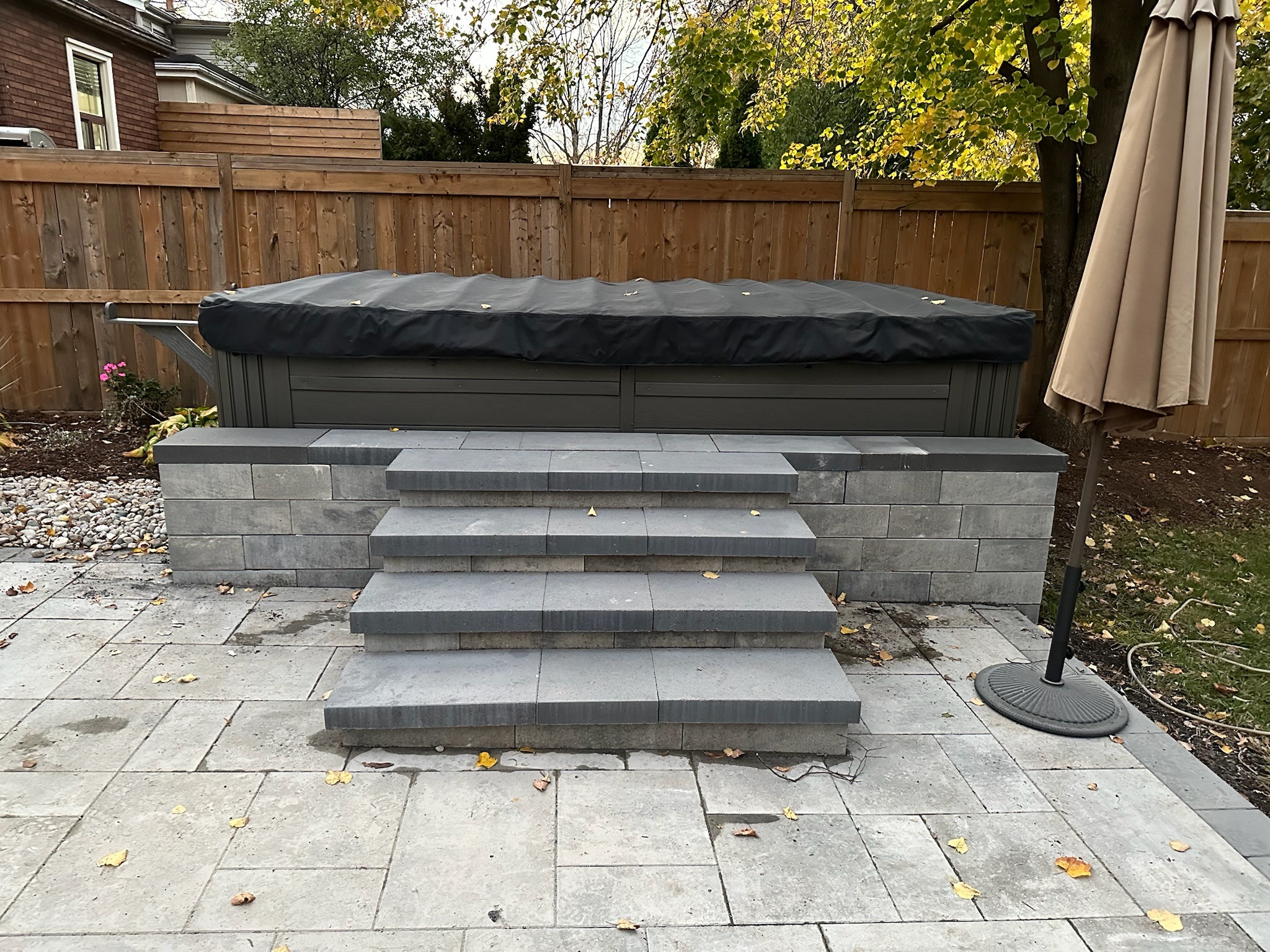 A backyard with a covered hot tub, stone steps, patio, wooden fence, and a closed umbrella, surrounded by trees and foliage.