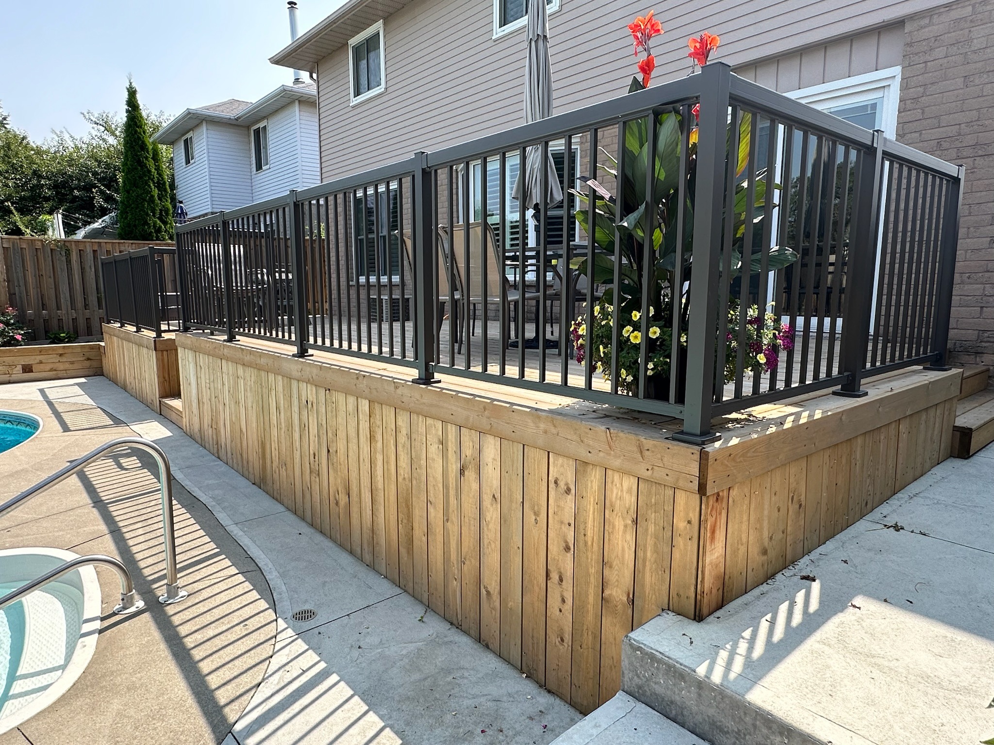 A wooden patio with black railing overlooks a backyard pool, adorned with vibrant flowers and outdoor seating, adjacent to a modern house.