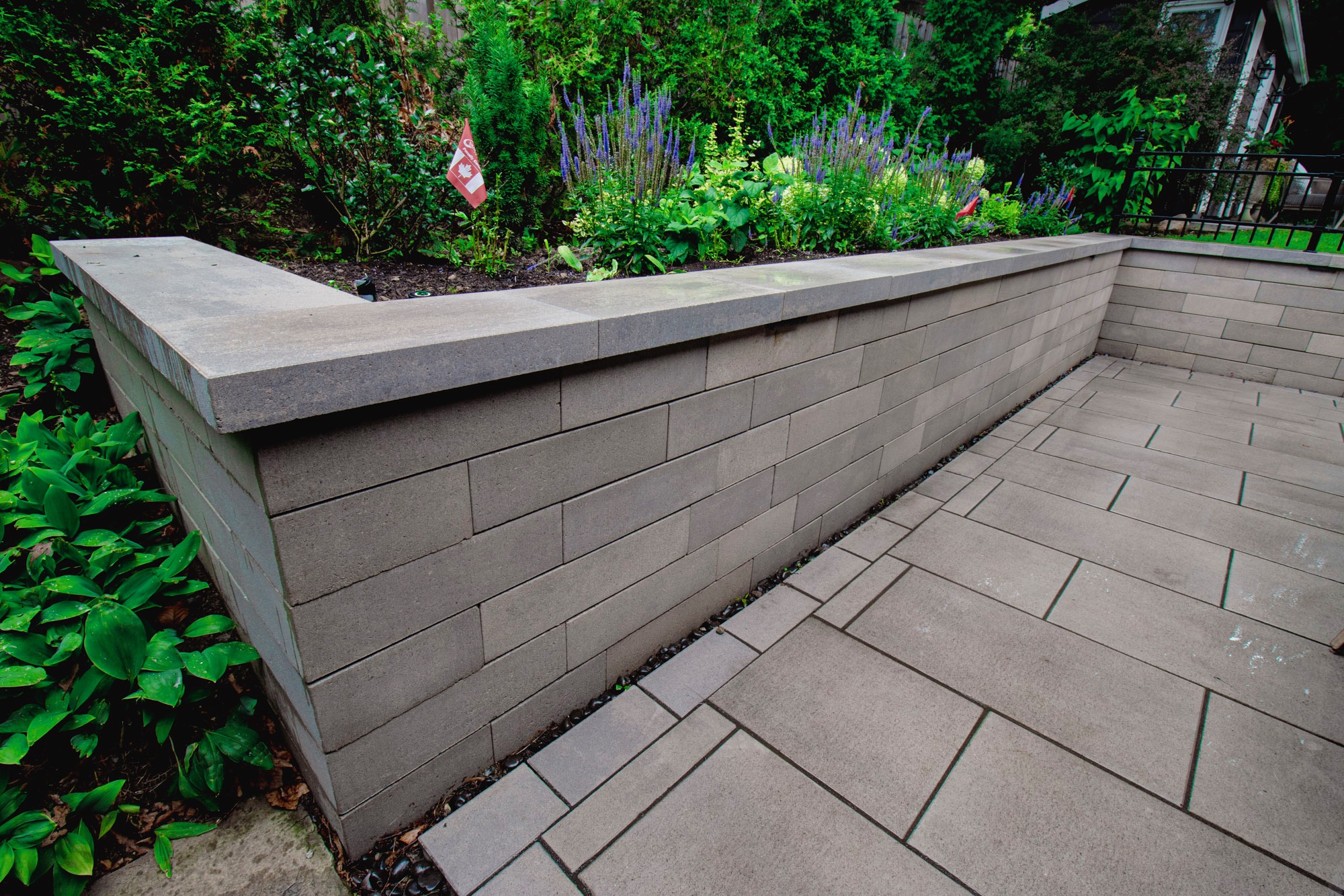 A patio with gray pavers and stone wall, bordered by lush green garden. Small Canadian flag in planter. Quiet, inviting outdoor space.