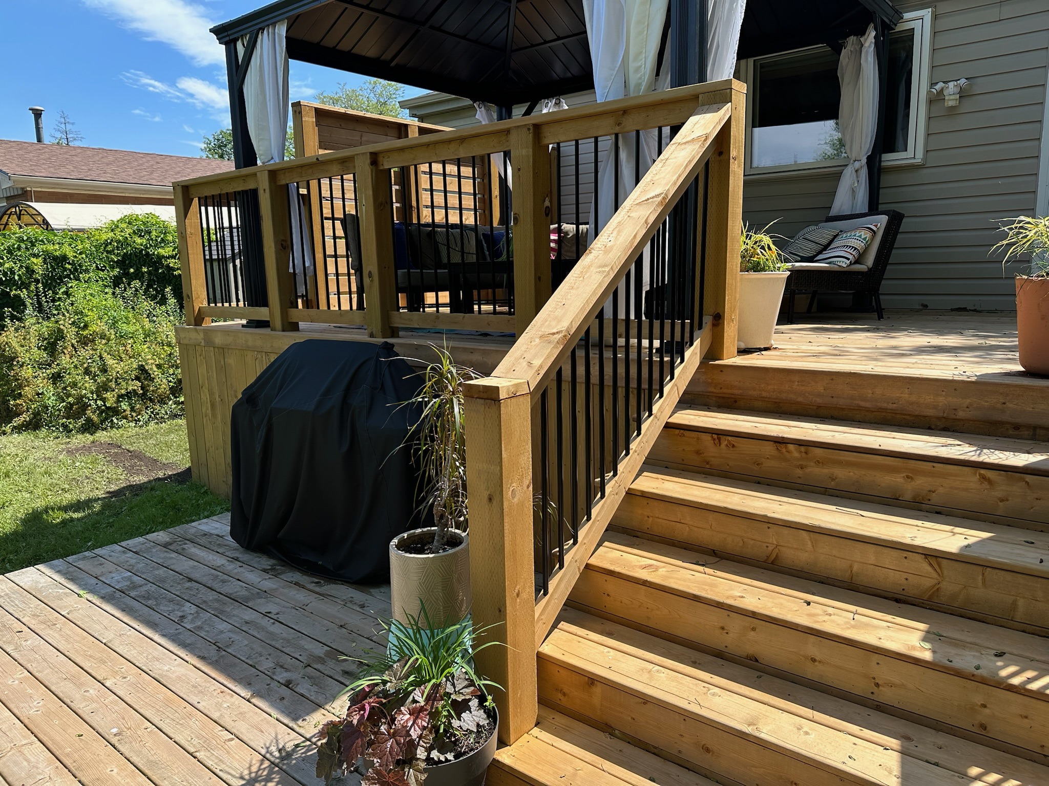 Wooden deck with stairs leading to a covered seating area. Potted plants decorate the foreground, surrounded by lush greenery under a clear sky.