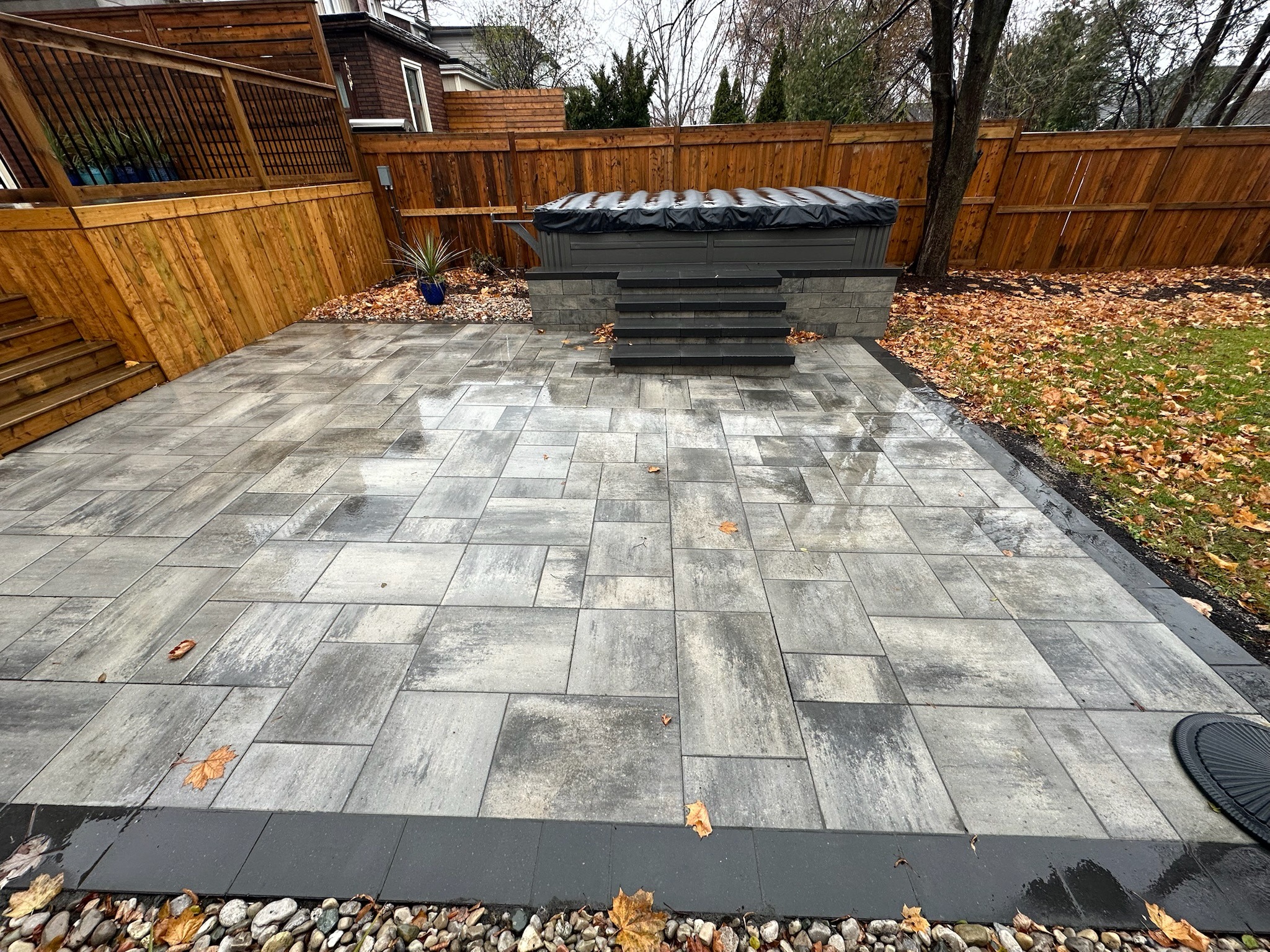 Spacious backyard patio with gray pavers, wooden fence, and covered hot tub. Autumn leaves scattered around, with trees and plants in the background.