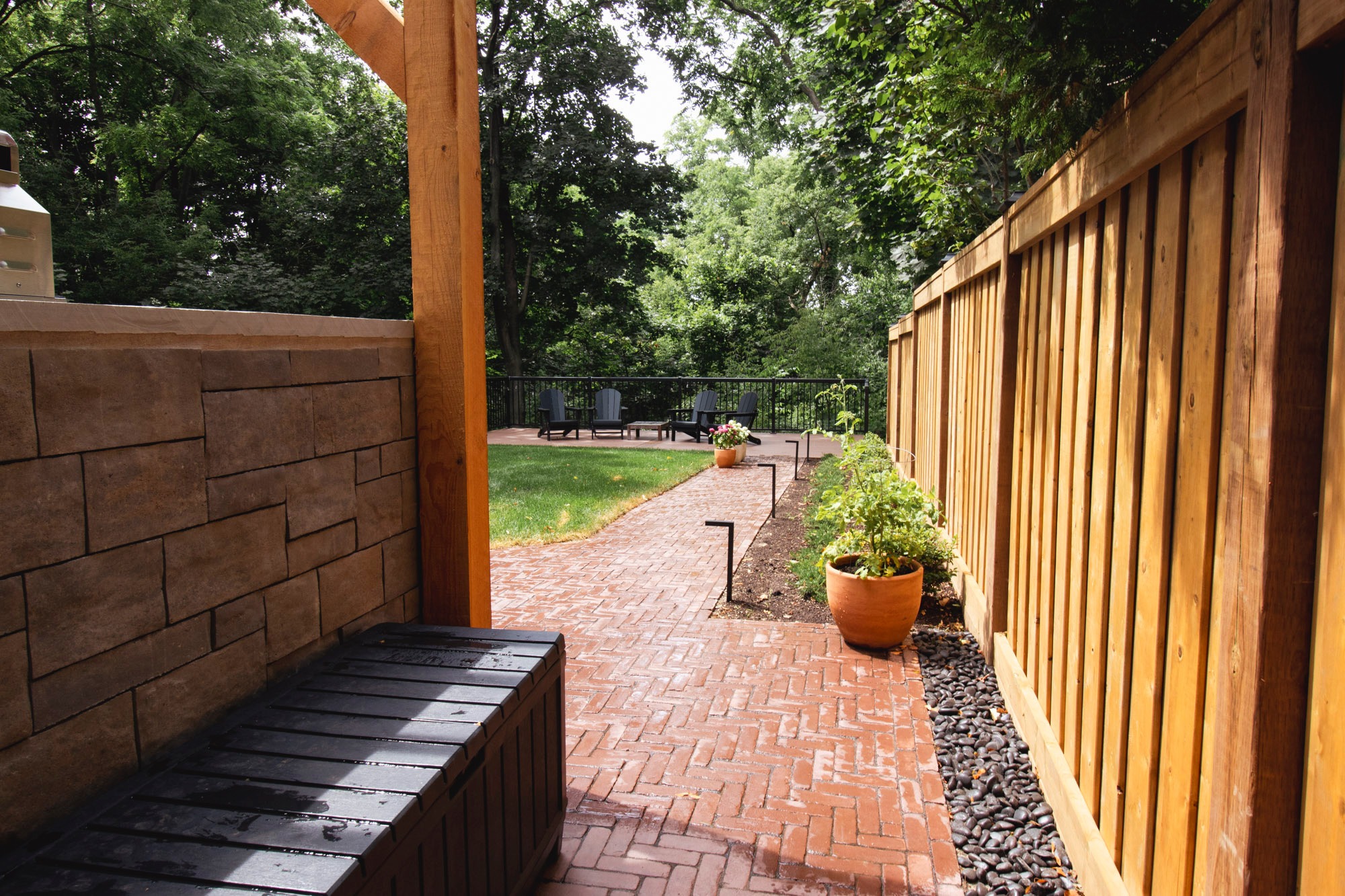A peaceful backyard features a brick patio, potted plants, wooden fencing, and outdoor seating surrounded by lush trees and greenery.