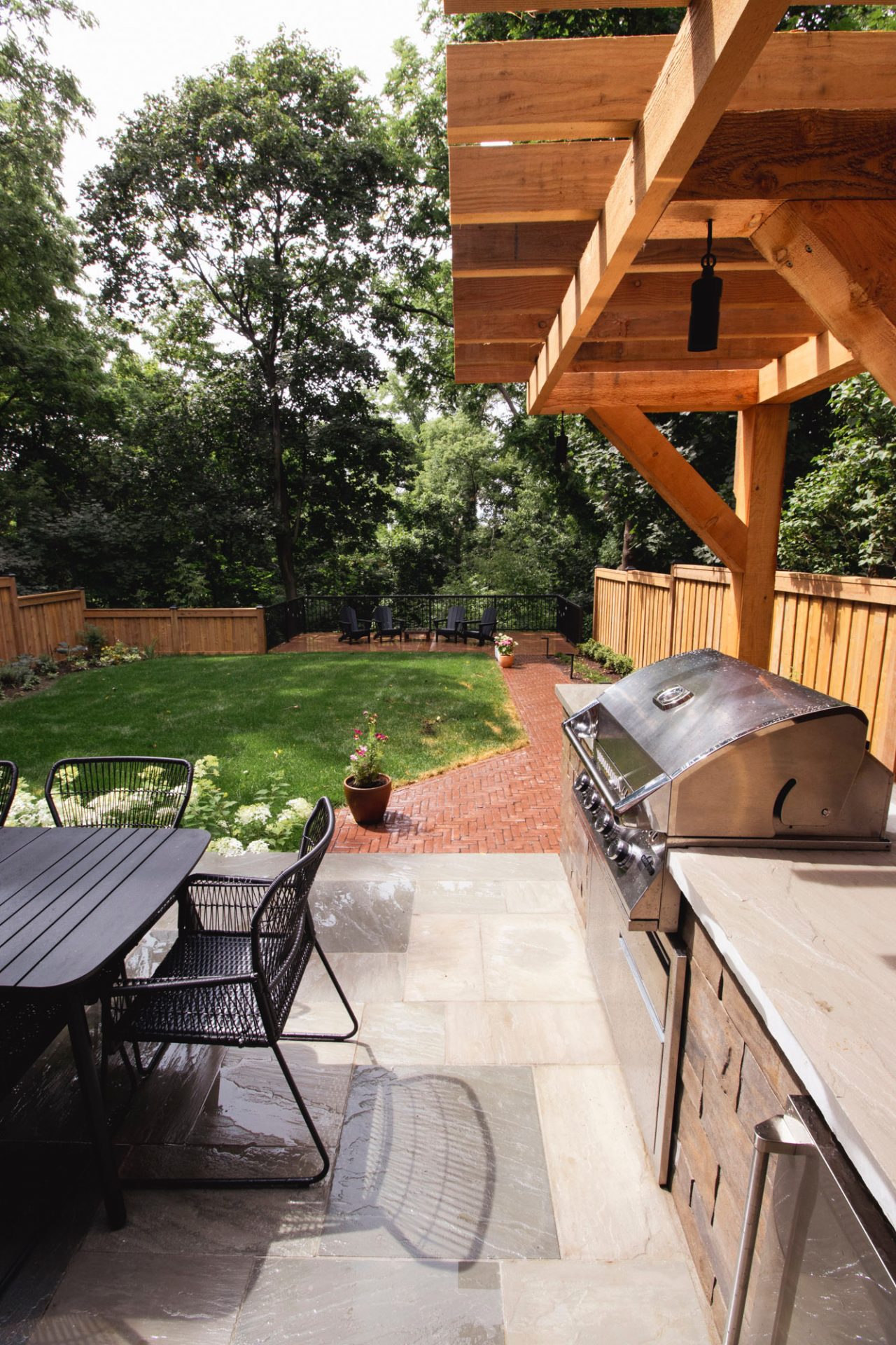 A wooden pergola covers a patio with a grill and seating. Brick path leads to more chairs in a landscaped backyard.