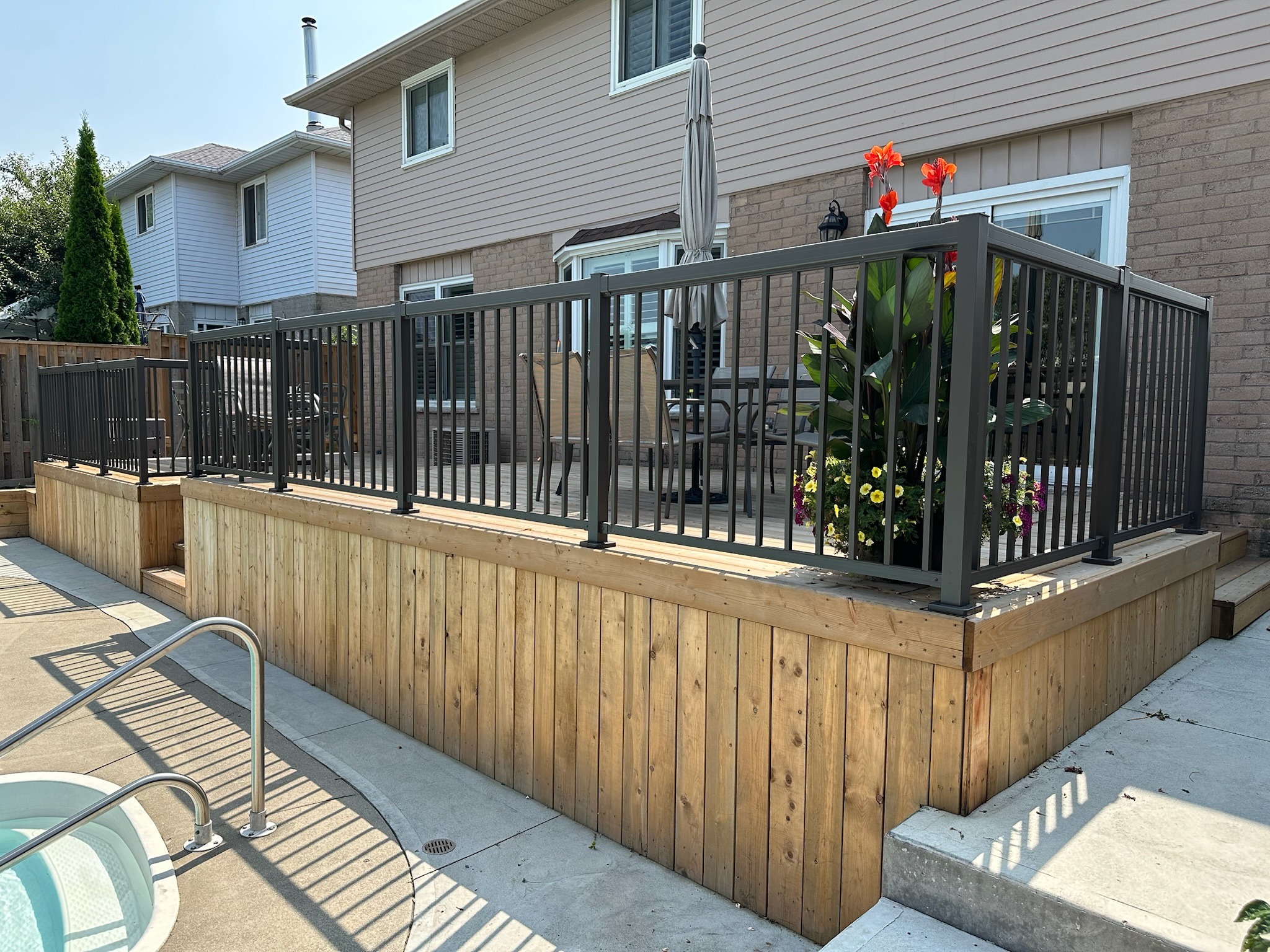 A backyard with a wooden deck, black railings, potted flowers, and a poolside area. Umbrella and patio furniture are visible.
