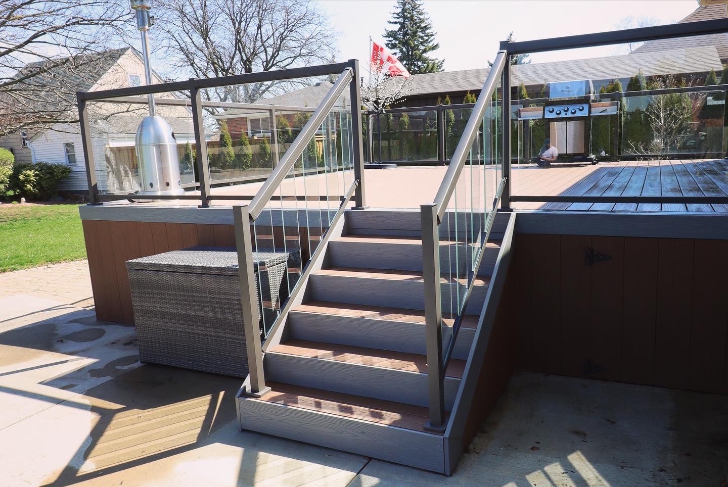 Modern backyard deck with glass railings and metal stairs, barbecue grill, wicker storage box, and a Canadian flag in the background.