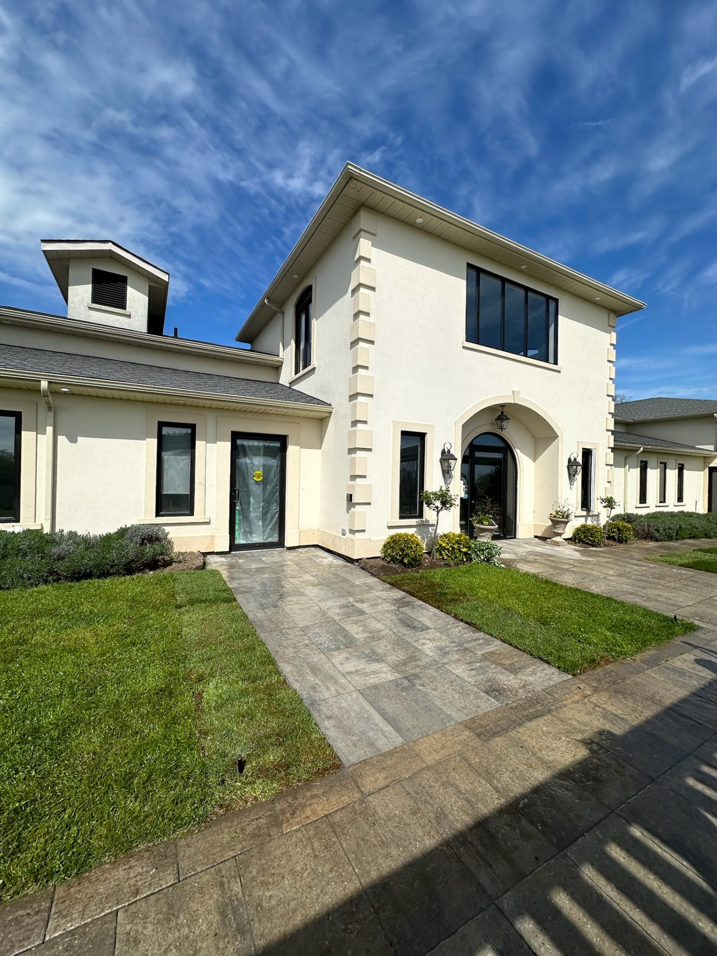 Modern two-story cream-colored house with large windows, lush green lawn, and blue sky. Paved walkway leads to entrance; no persons visible.