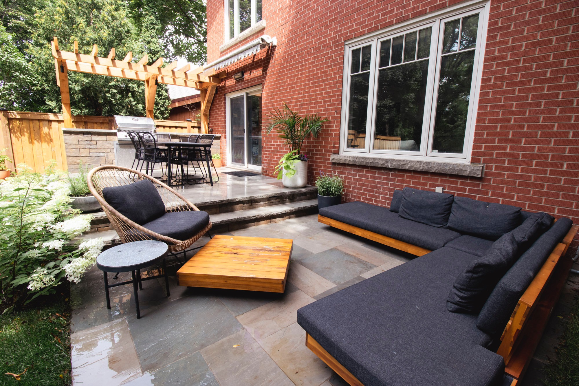 A brick house features a cozy outdoor patio with modern seating, pergola, grill, dining area, lush plants, and stone flooring.