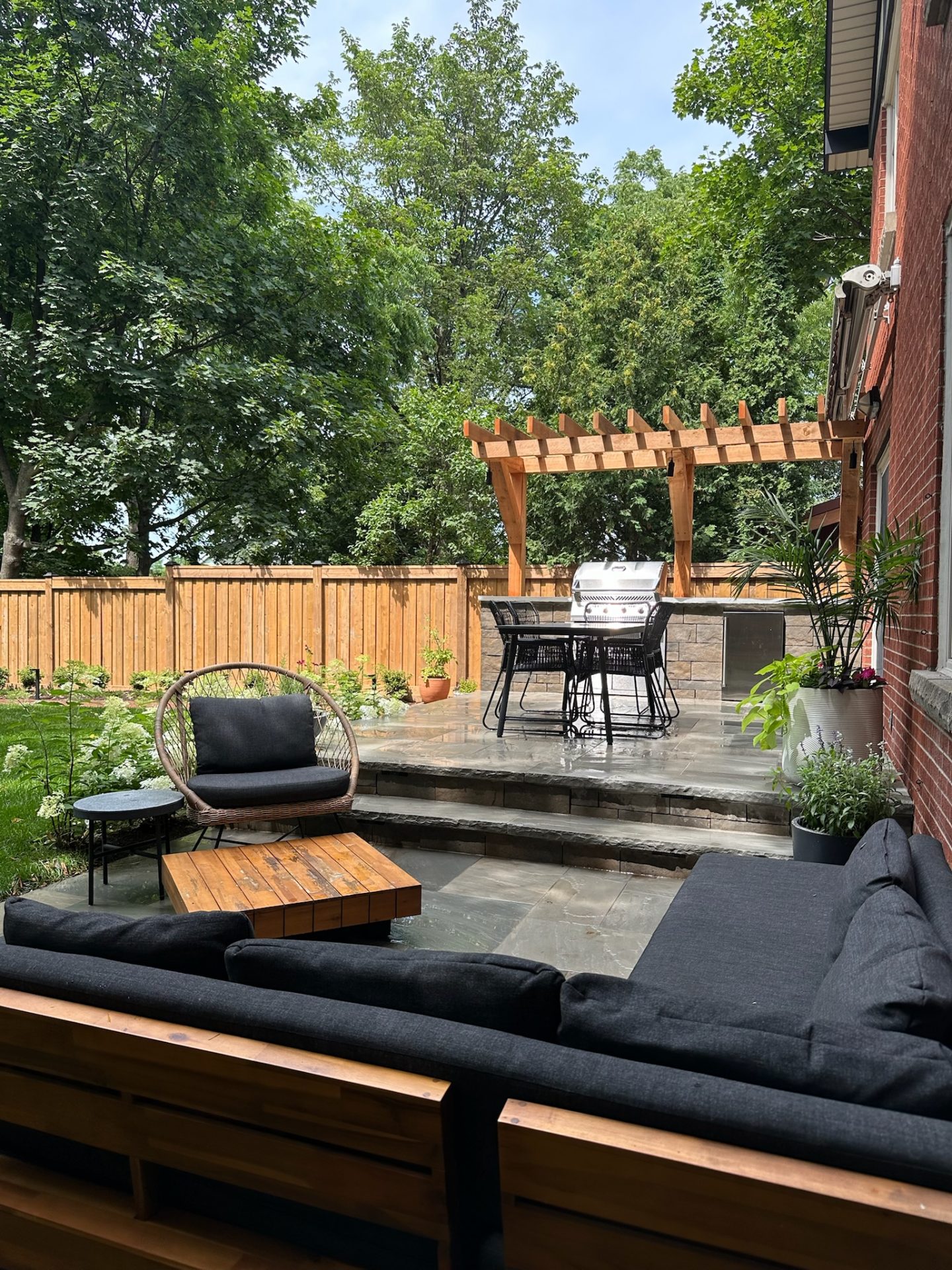 A cozy backyard patio features wooden furniture, a pergola, and a barbecue grill. Surrounded by greenery and a wooden fence, perfect for relaxation.
