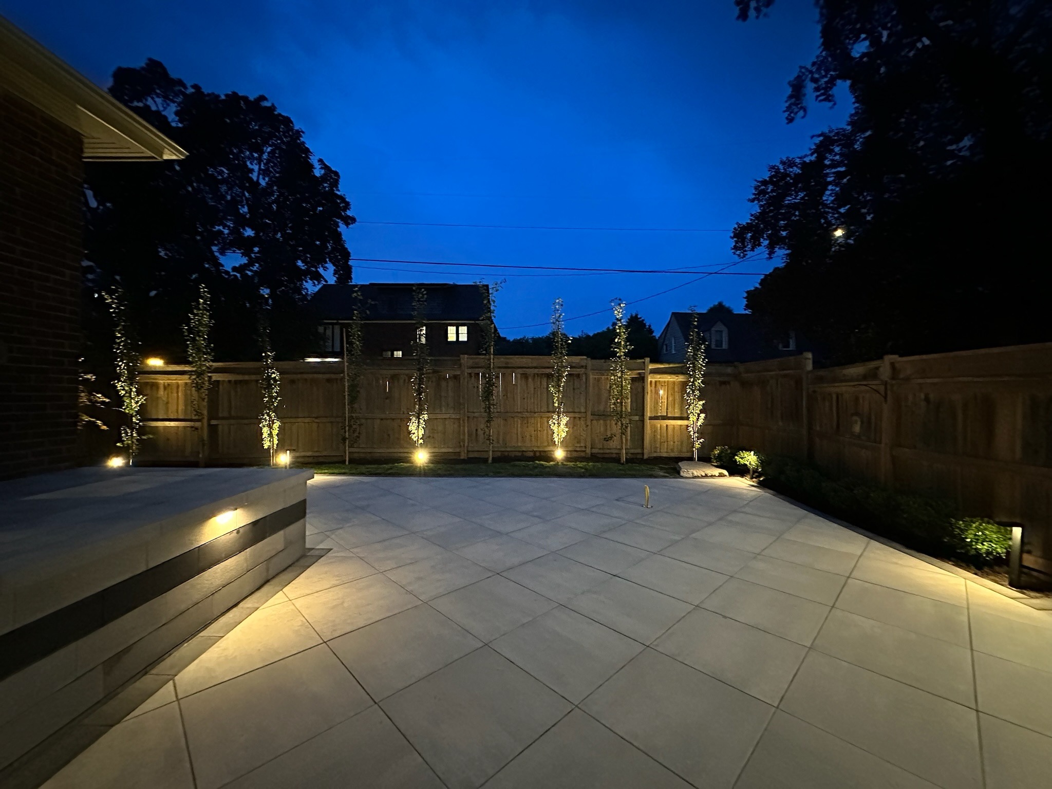 A well-lit backyard at night with patio tiles, plants, and wooden fence, surrounded by trees. There's a modern exterior house view.