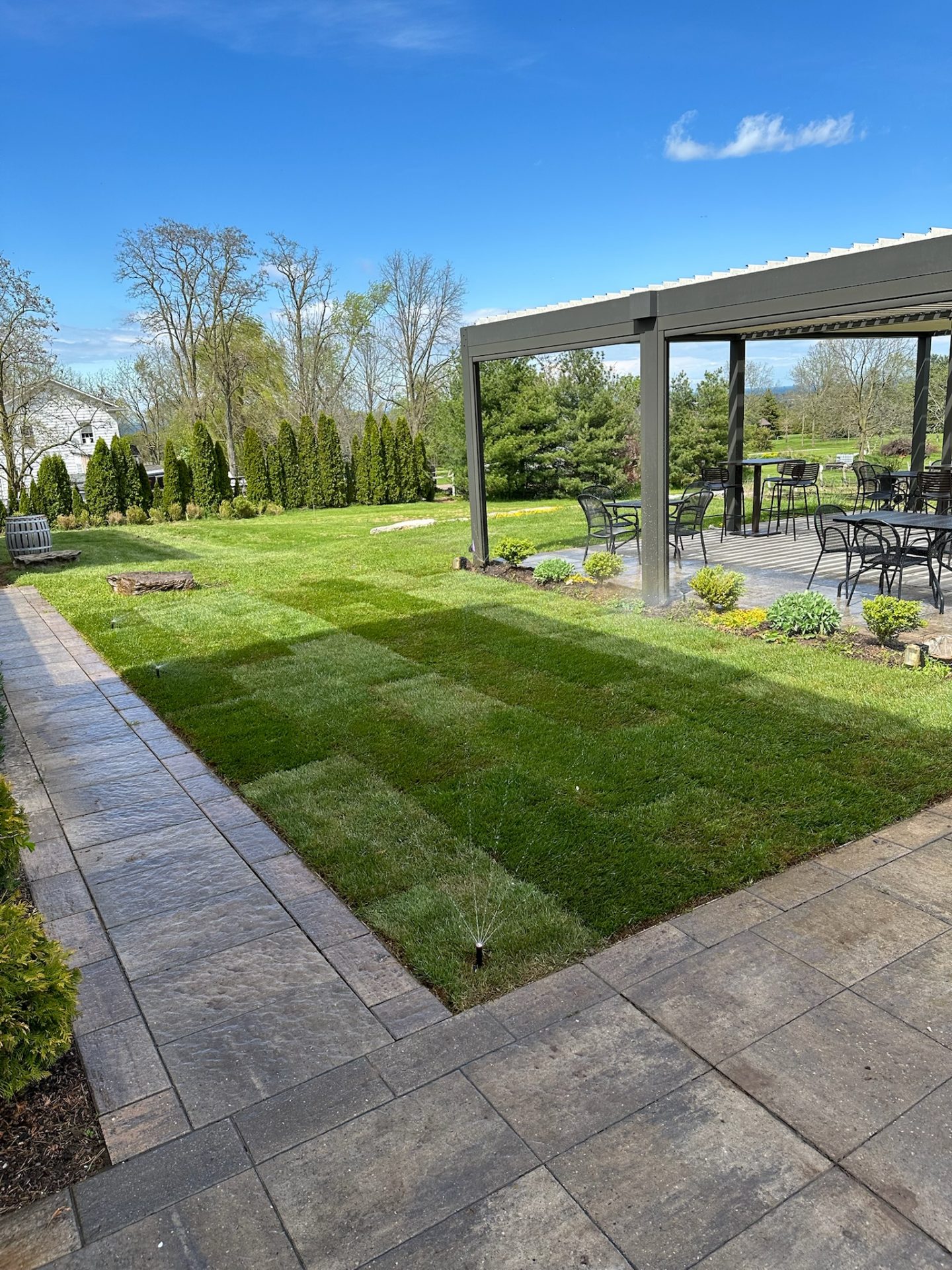 A well-maintained garden features a paved path, sprinkler, lush grass, and a covered patio with tables and chairs under a clear blue sky.
