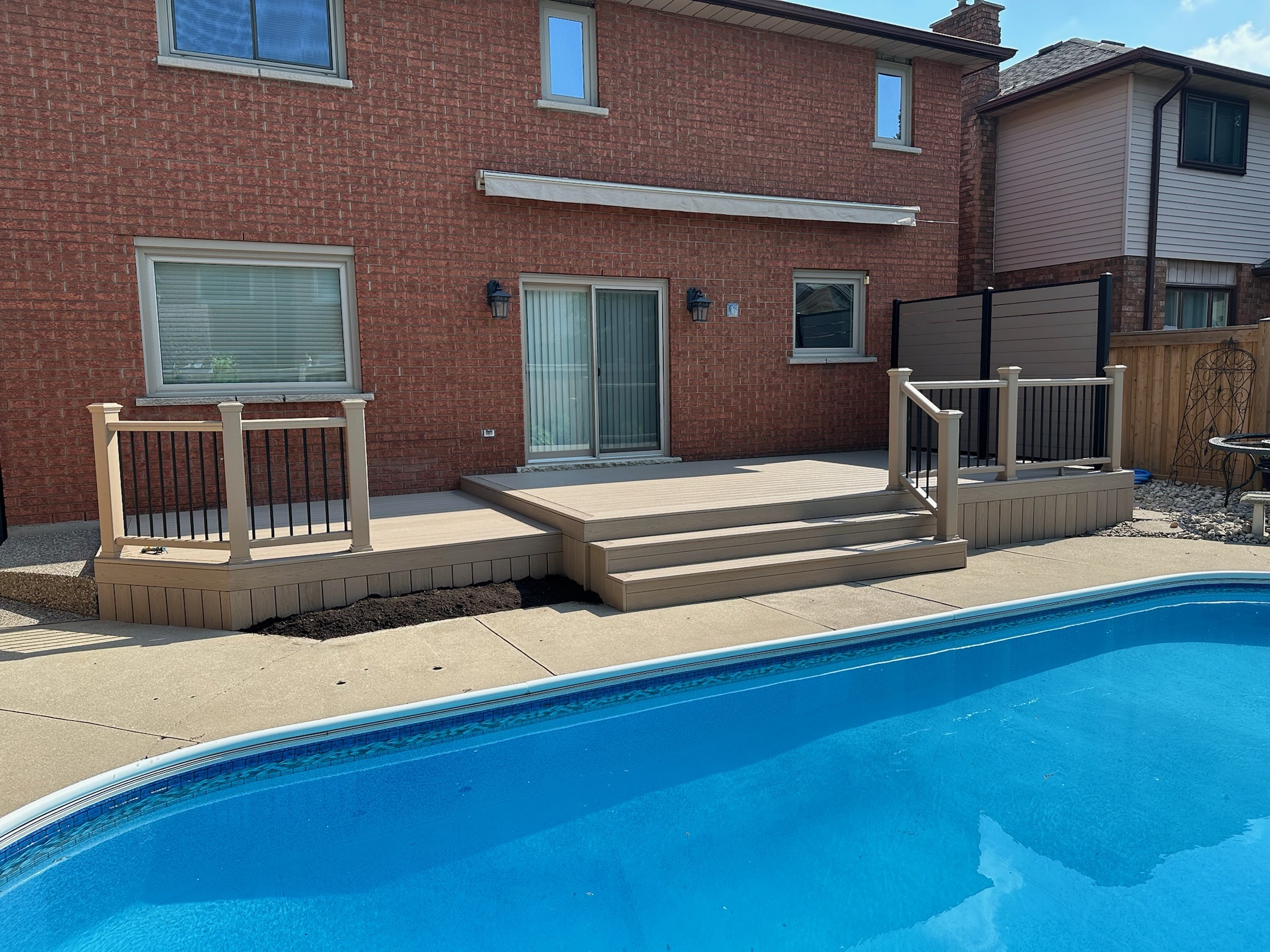 Brick house with a wooden deck and steps leading to an adjacent swimming pool. Clear, sunny day with no people visible.
