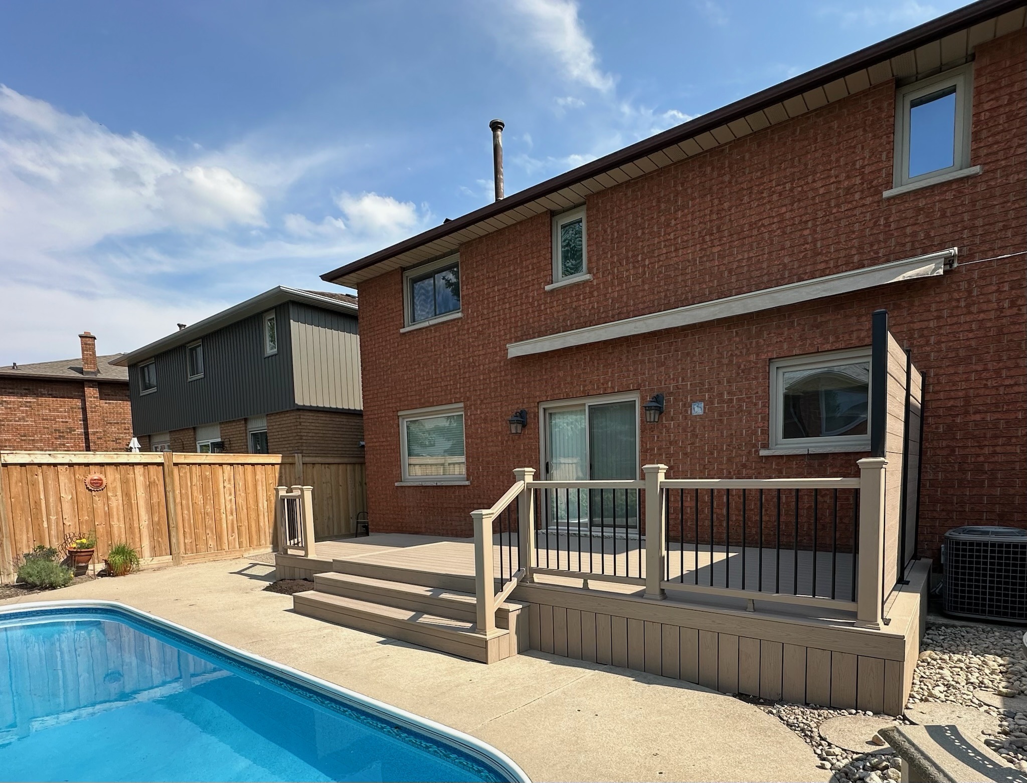 Brick house with a wooden deck overlooks a clear swimming pool. Sunny day with blue sky, surrounded by a wooden fence and neighboring homes.