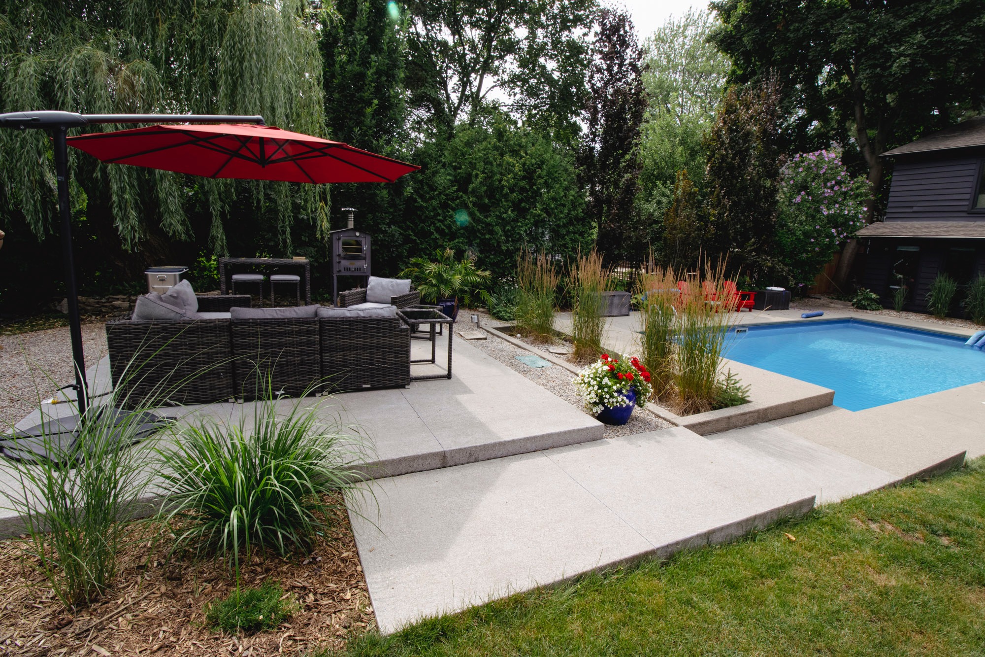 A backyard patio with a pool, surrounded by greenery, features outdoor seating under a red umbrella, and vibrant potted plants.