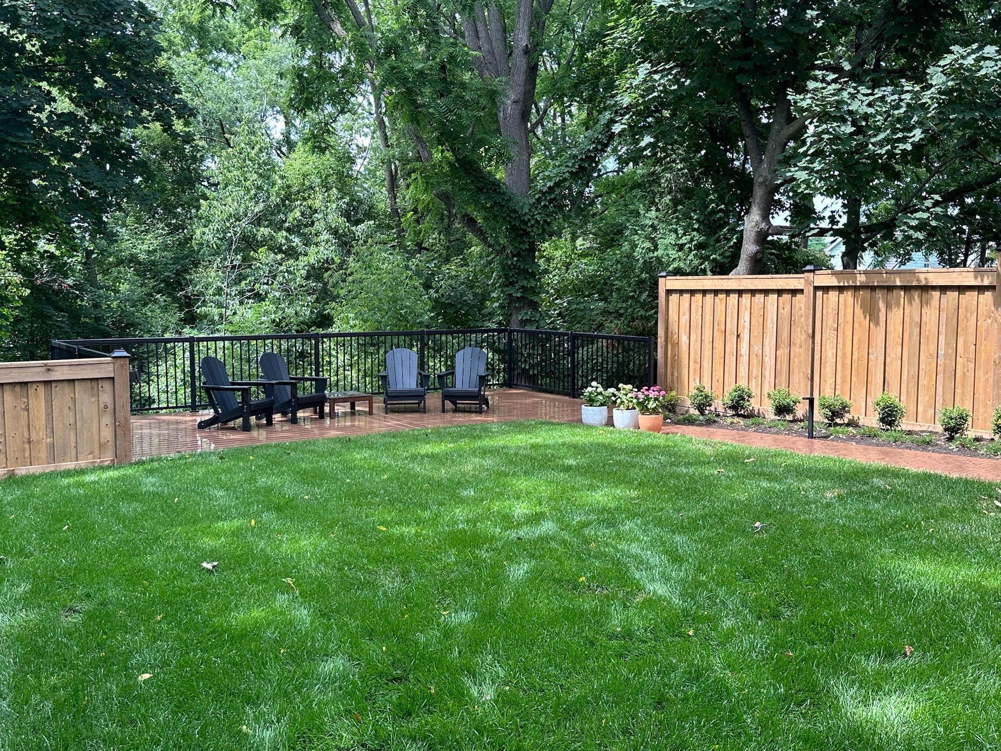 A lush backyard features Adirondack chairs on a wooden deck, surrounded by vibrant greenery and a wooden fence with potted flowers.
