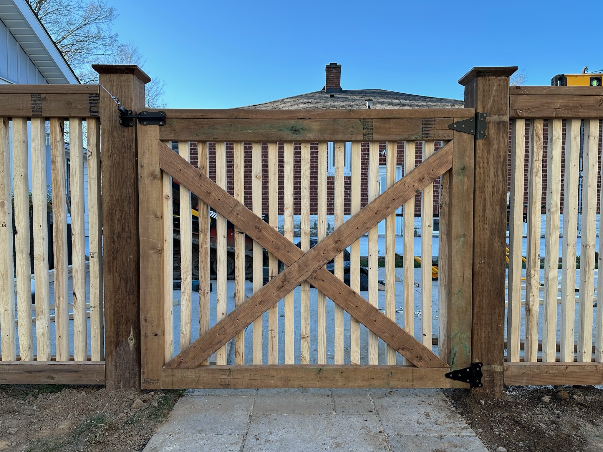 Wooden gate with diagonal braces and vertical slats, bordered by sturdy posts, stands in a backyard with clear blue sky backdrop.