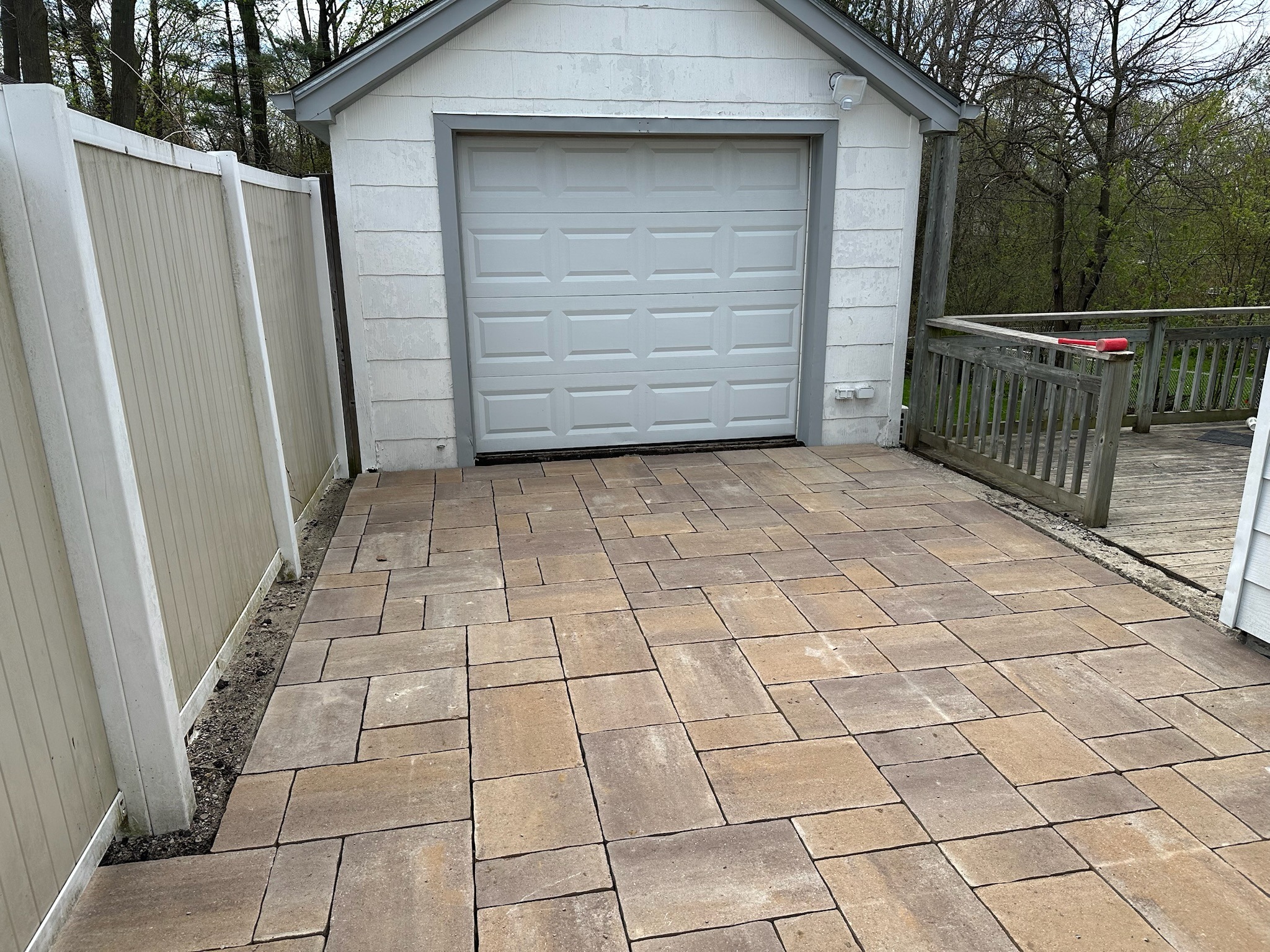 A small shed with a closed garage door stands, surrounded by beige fencing and a stone-paved driveway, amidst trees.