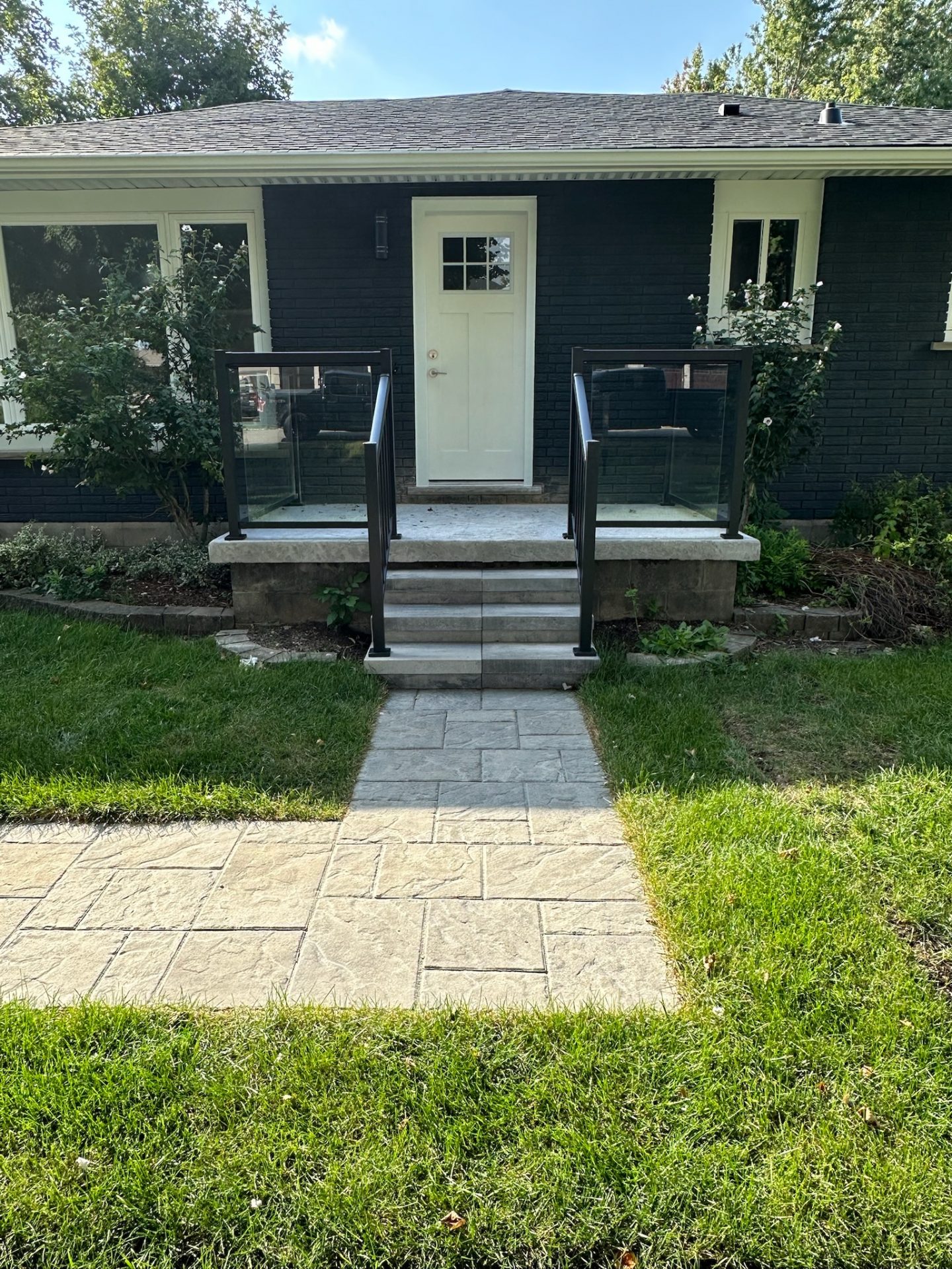A modern house with a black brick exterior, white door, and glass railings. Paved walkway leads through well-maintained lawn and garden.